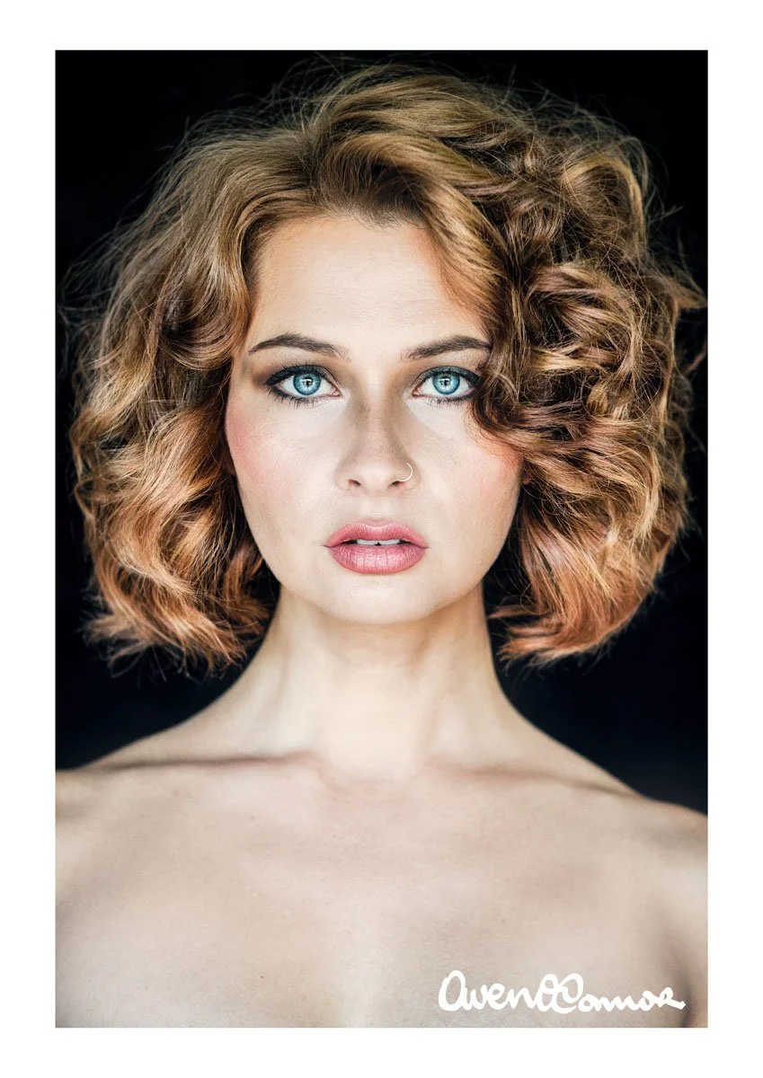 Portrait of a woman with curly, shoulder-length auburn hair and blue eyes, looking directly at the camera against a black background, with a signature 'AwenOComa' at the bottom.