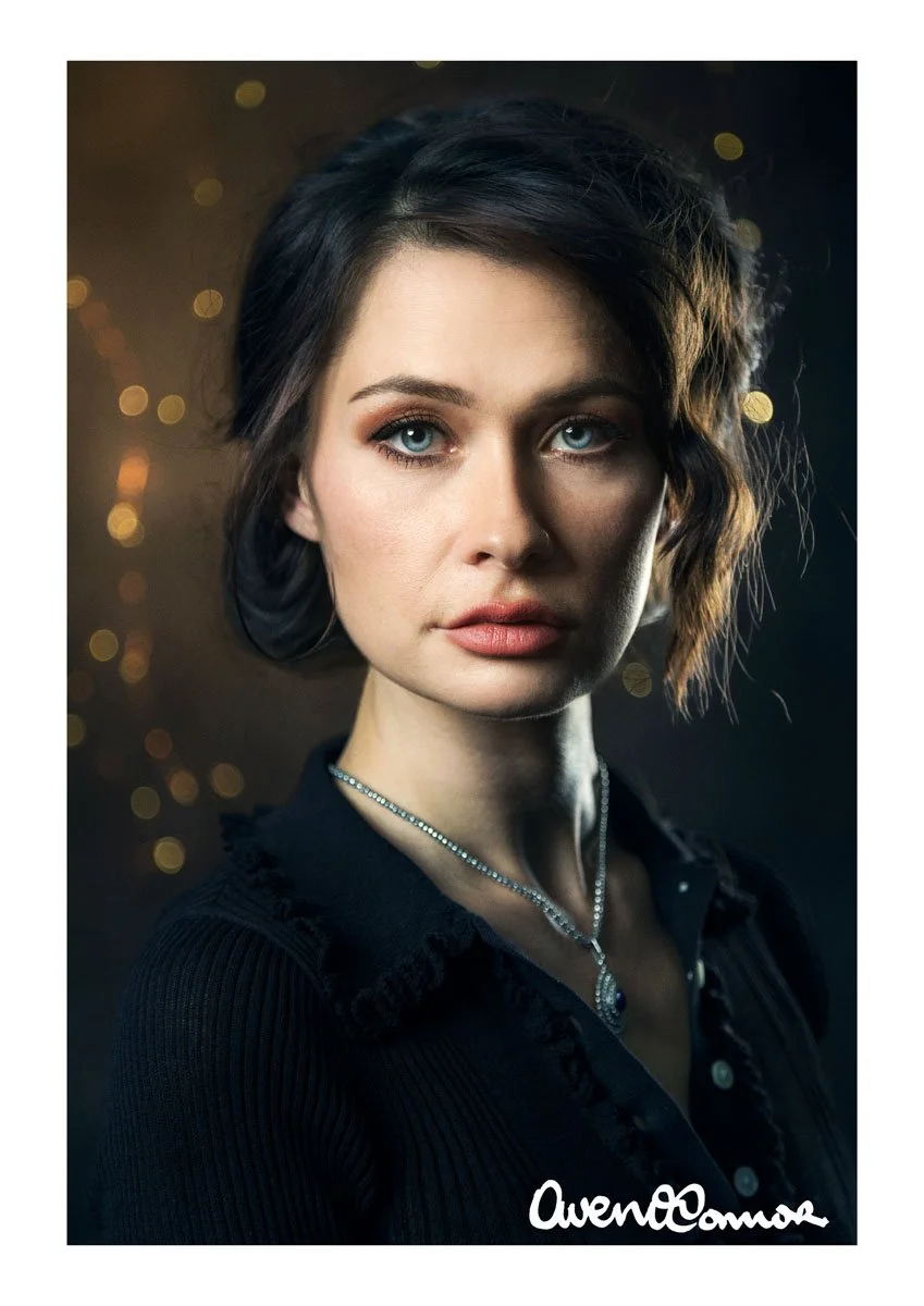 A woman with fair skin, short wavy dark brown hair, blue eyes, wearing makeup and a black top with ruffled lace details, a diamond necklace, and a pendant. She has a serious expression and is looking directly at the camera with a dark, blurred backgr