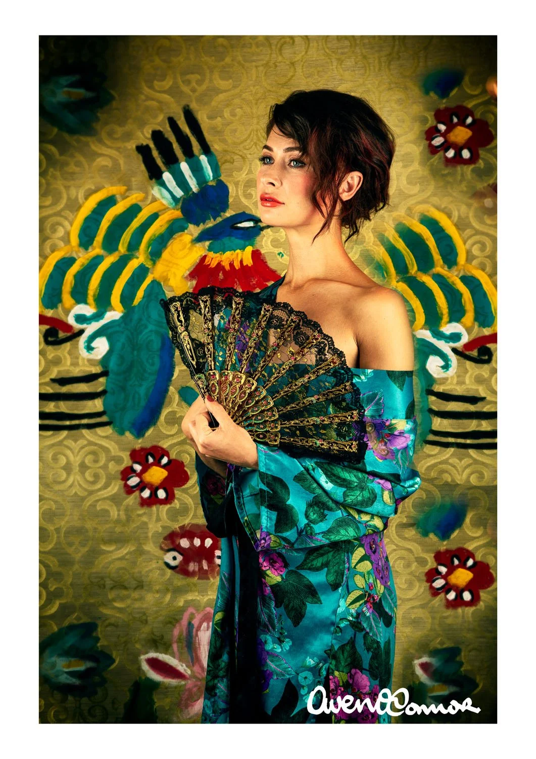 Woman with short dark hair holding a decorative fan, wearing a floral blue dress, behind a colorful painted background.