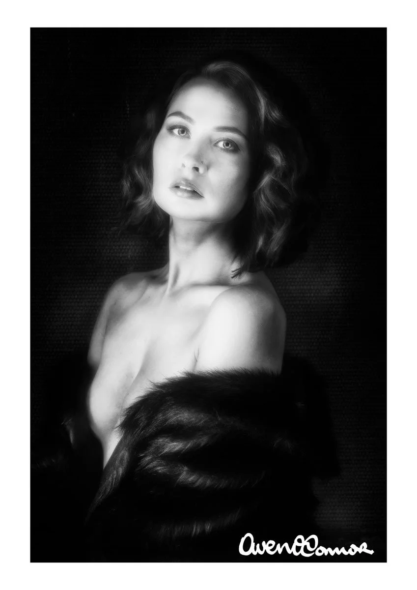Black and white portrait of a woman with wavy hair, looking directly at the camera, partially covered with a fur garment, signed 'OwenComas' in the bottom right corner.