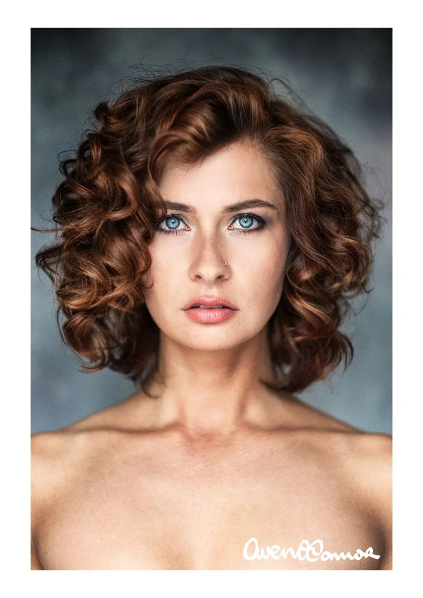 A portrait of a woman with curly auburn hair and blue eyes, looking directly at the camera against a dark gray background.