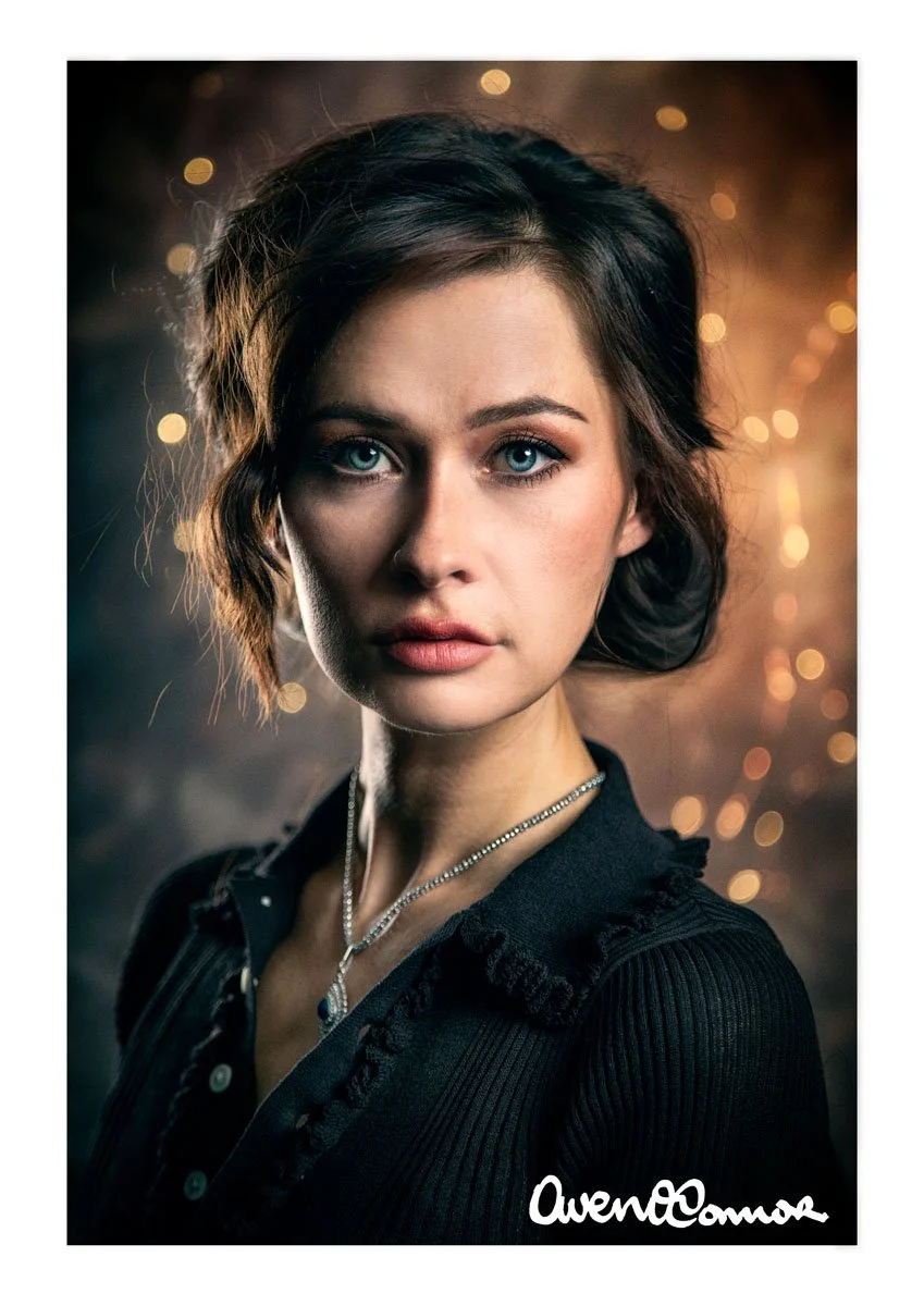 A young woman with short, wavy brown hair and blue eyes wearing a black shirt and a silver necklace, posed against a blurred background with warm light bokeh effects.