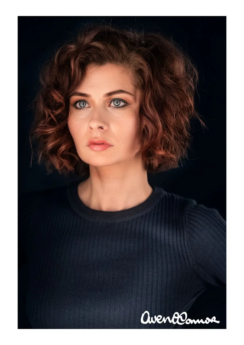 A woman with curly red hair and blue eyes wearing a black top, looking to the side against a dark background.
