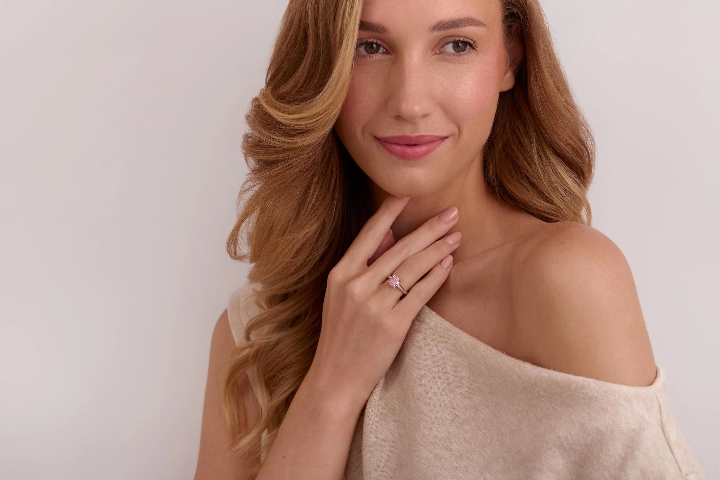A woman with long wavy red hair and hazel eyes, smiling softly. She is touching her chin with her hand, which is adorned with a ring. She is wearing a beige sleeveless top and standing against a plain light background. Jewellry on model photography