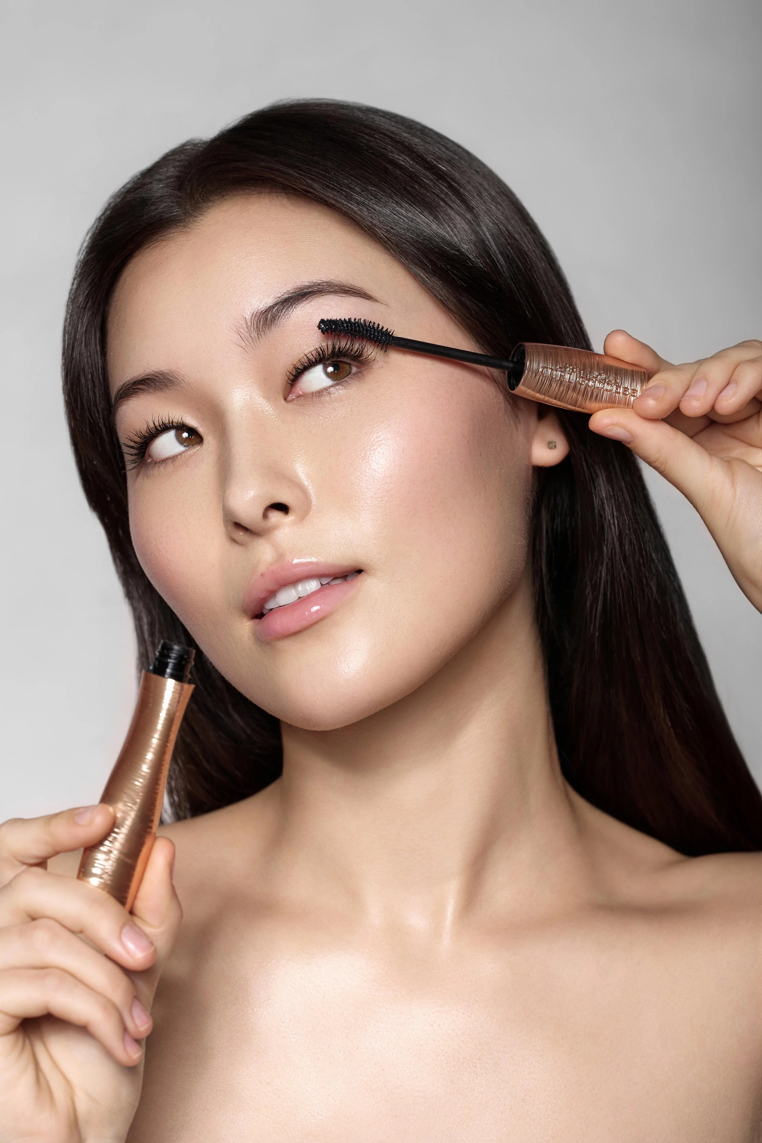 A model with dark brown hair applying mascara by Lancome to her eyelashes, holding the mascara wand in her right hand and the mascara tube in her left hand. Beauty campaign for Lancome
