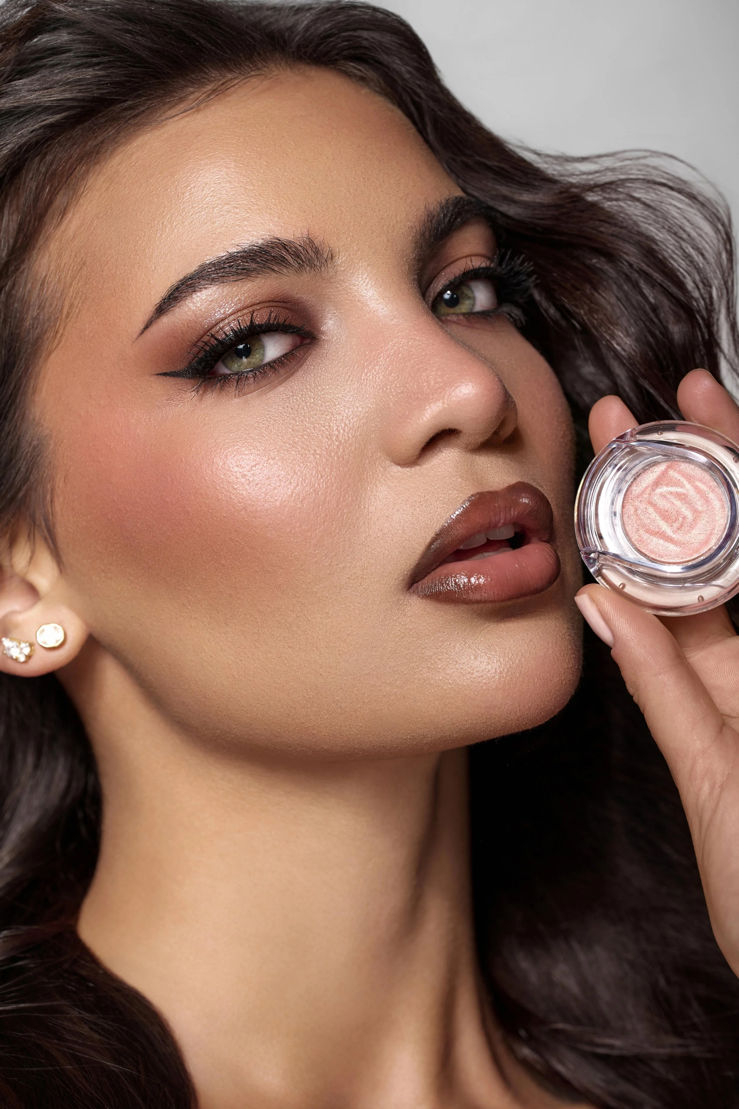 Close-up of a model with olive green eyes and tan skin holding a small container of shimmery pink cosmetic product near her lips. Beauty campaign for Lancome