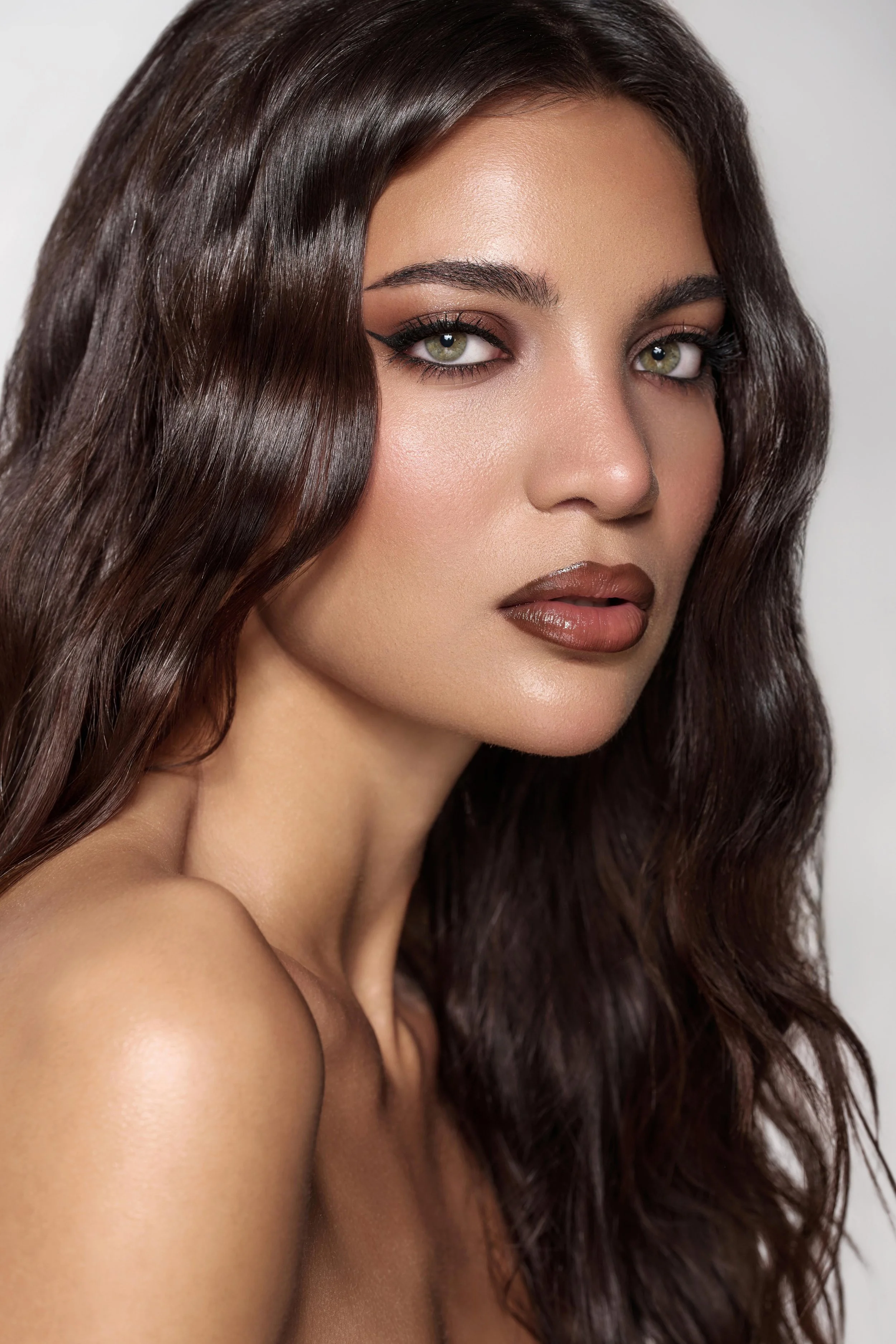 Close-up of a model with wavy dark brown hair, green eyes, and makeup including eyeliner and brown lipstick. Beauty portrait