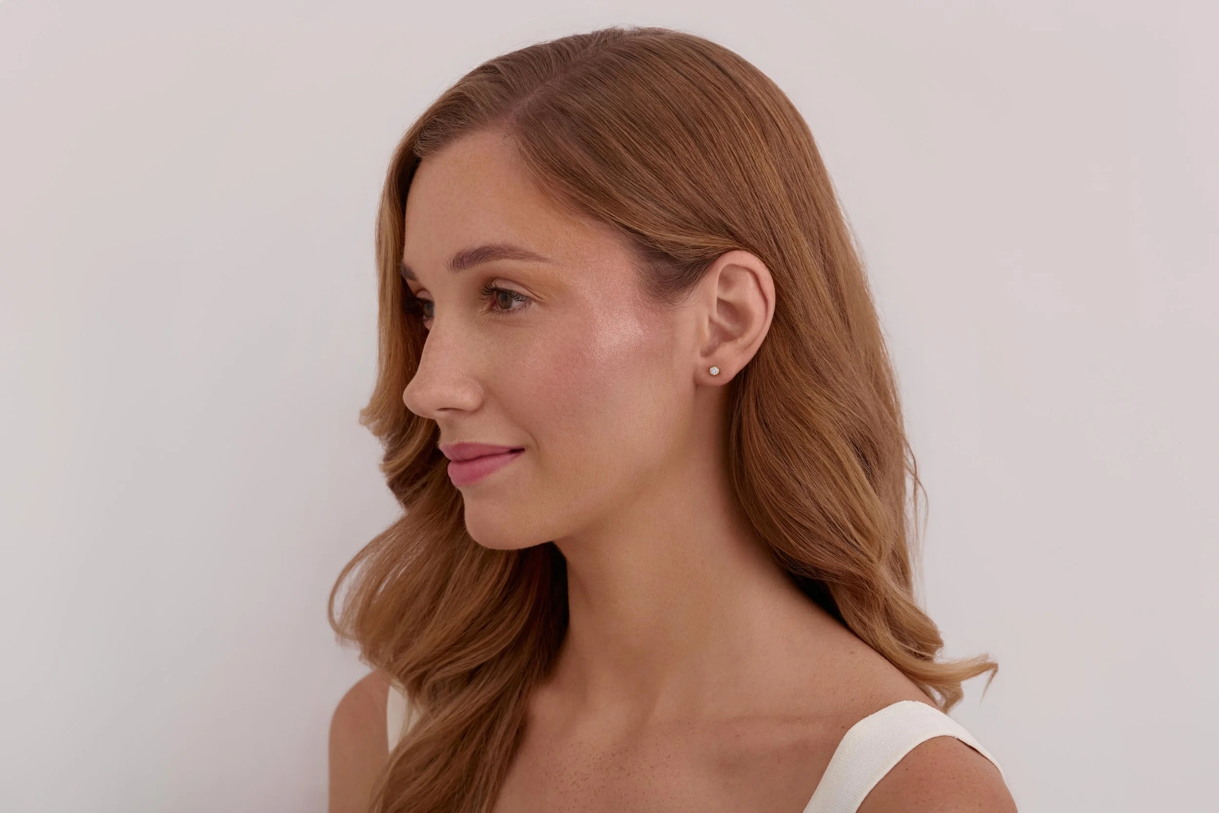 A woman with long red hair, styled in loose waves, wearing a white sleeveless top and small diamond stud earrings, looking to her left against a plain light-colored background. Jewellry on model photography in London