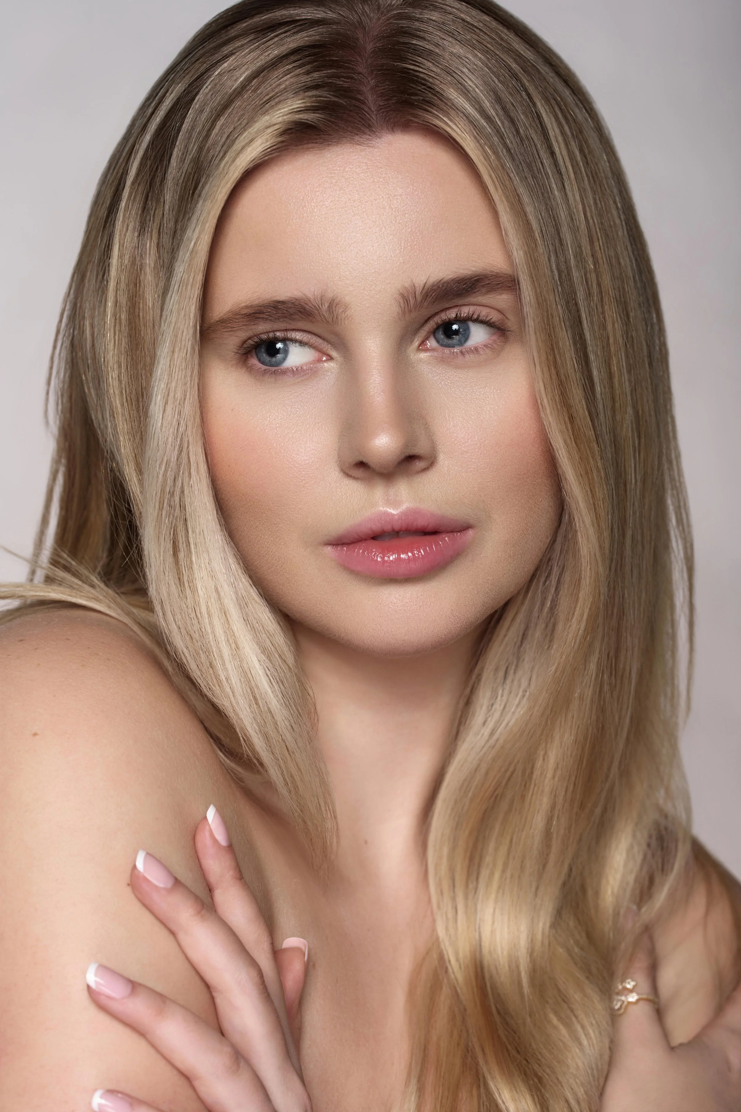A model with long blonde hair and blue eyes, posing with her hand on her shoulder, wearing pink lipstick and manicured nails. Model portfolio portrait