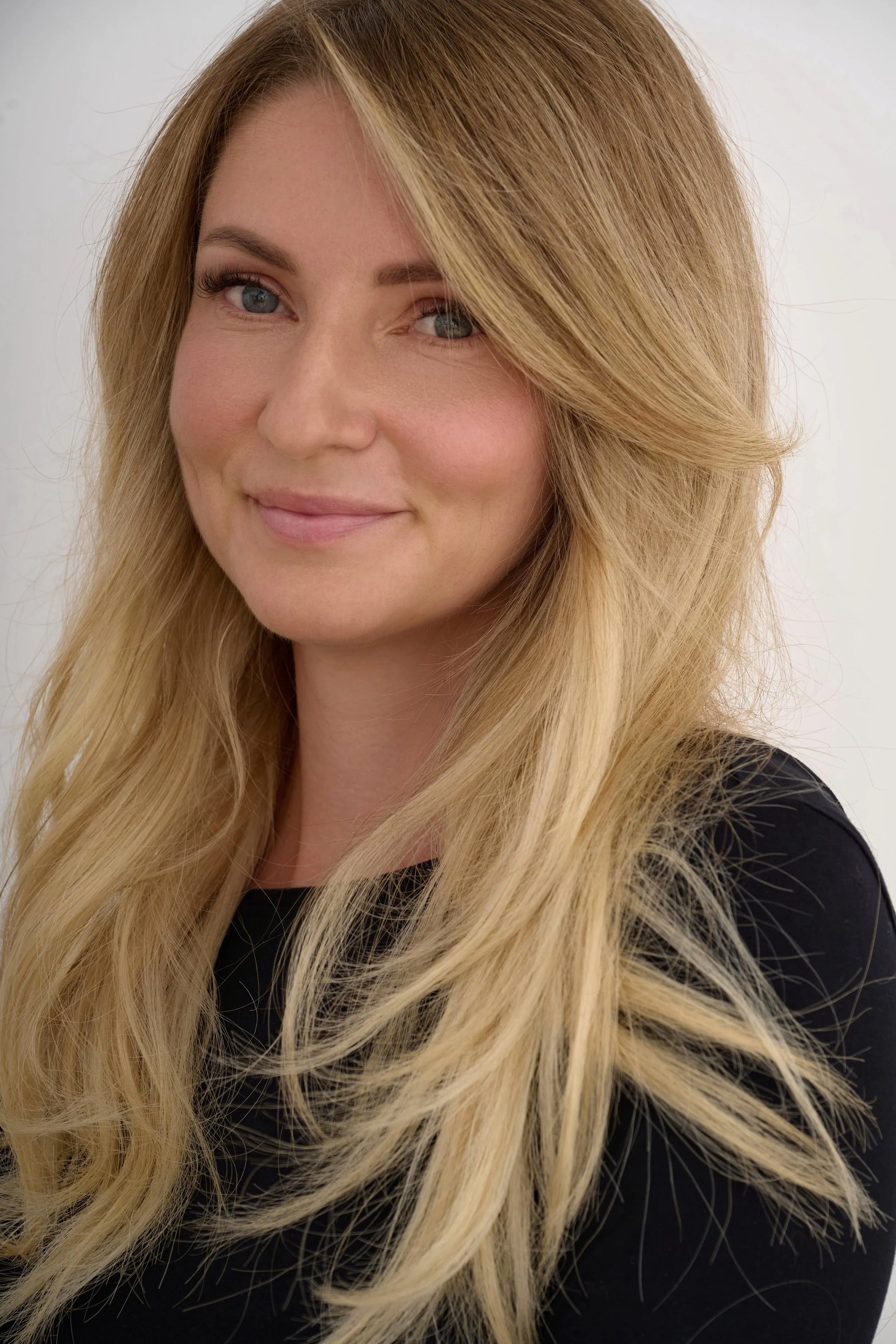 Close-up of a smiling Jana Kukebal, commercial photographer, with long blonde hair, wearing a black top, against a plain light background.