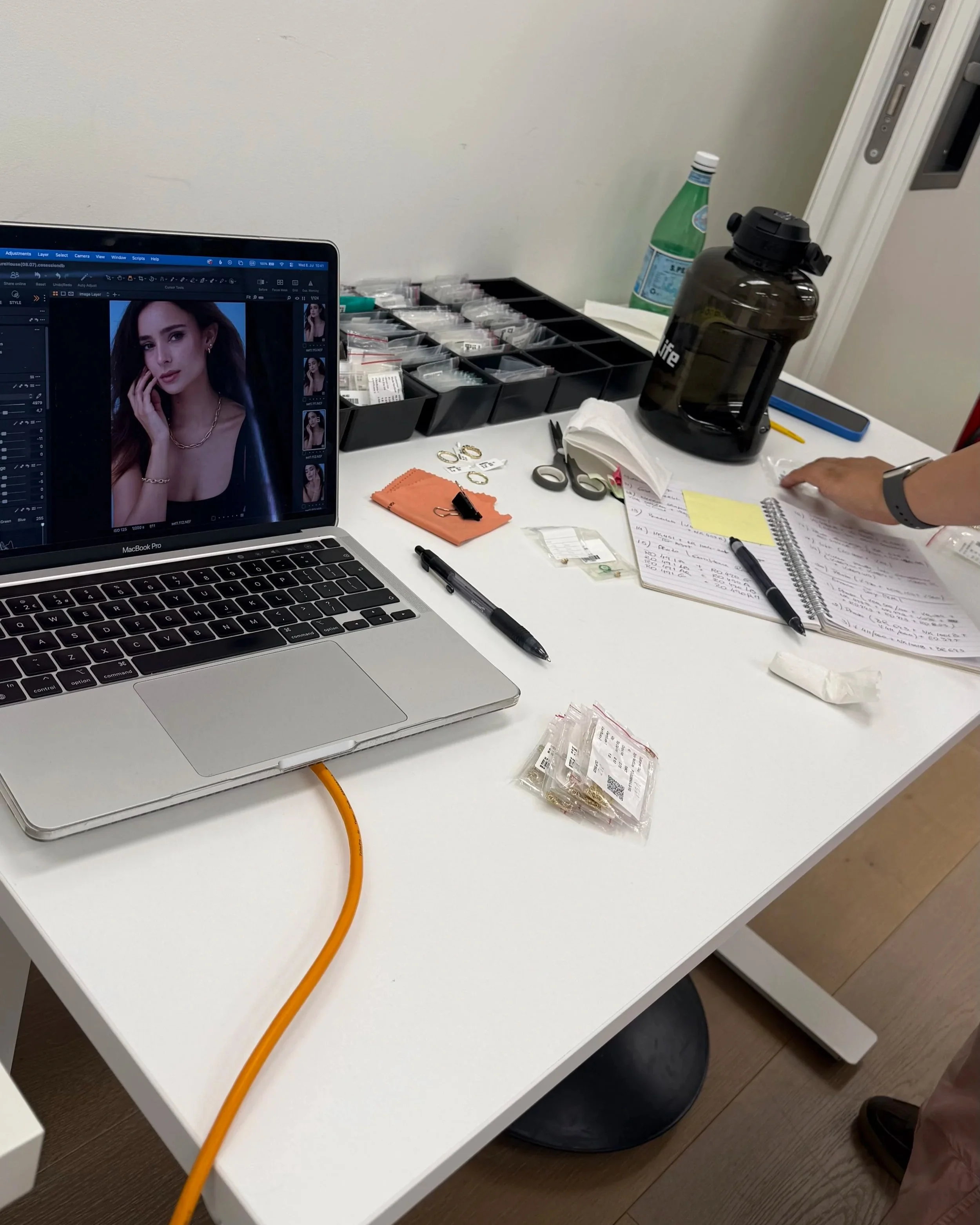 A photographer's workspace with a laptop showing a portrait photo editing software, small jewelry items, scissors, a water bottle, a black water container, notebooks, papers, a pen, and an orange extension cord on a white desk.