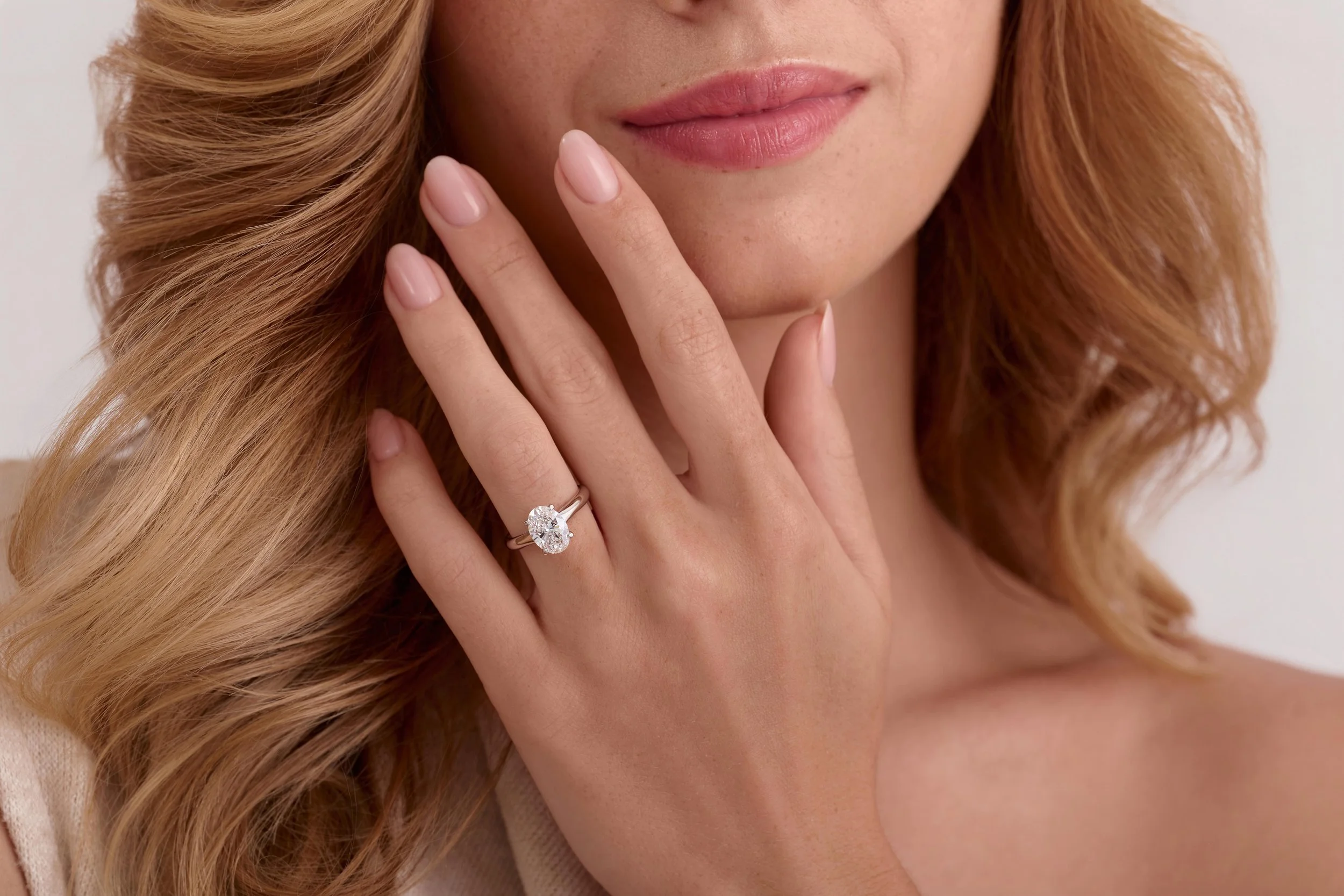 Close-up of a model with blonde wavy hair, pink lipstick, and a large diamond engagement ring on her finger, touching her face. Ecommerce photography for jewellery brand