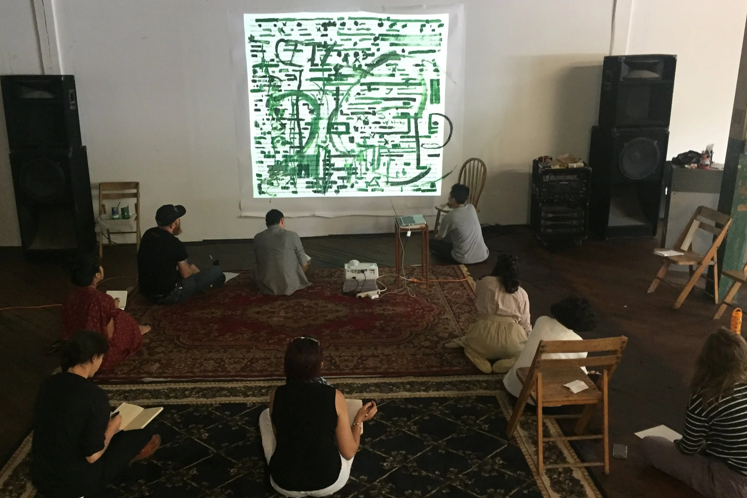 View of the room, with people sitting scattered around the projection of the poem, mostly painted over by green marks. The people are writing in individual note books.
