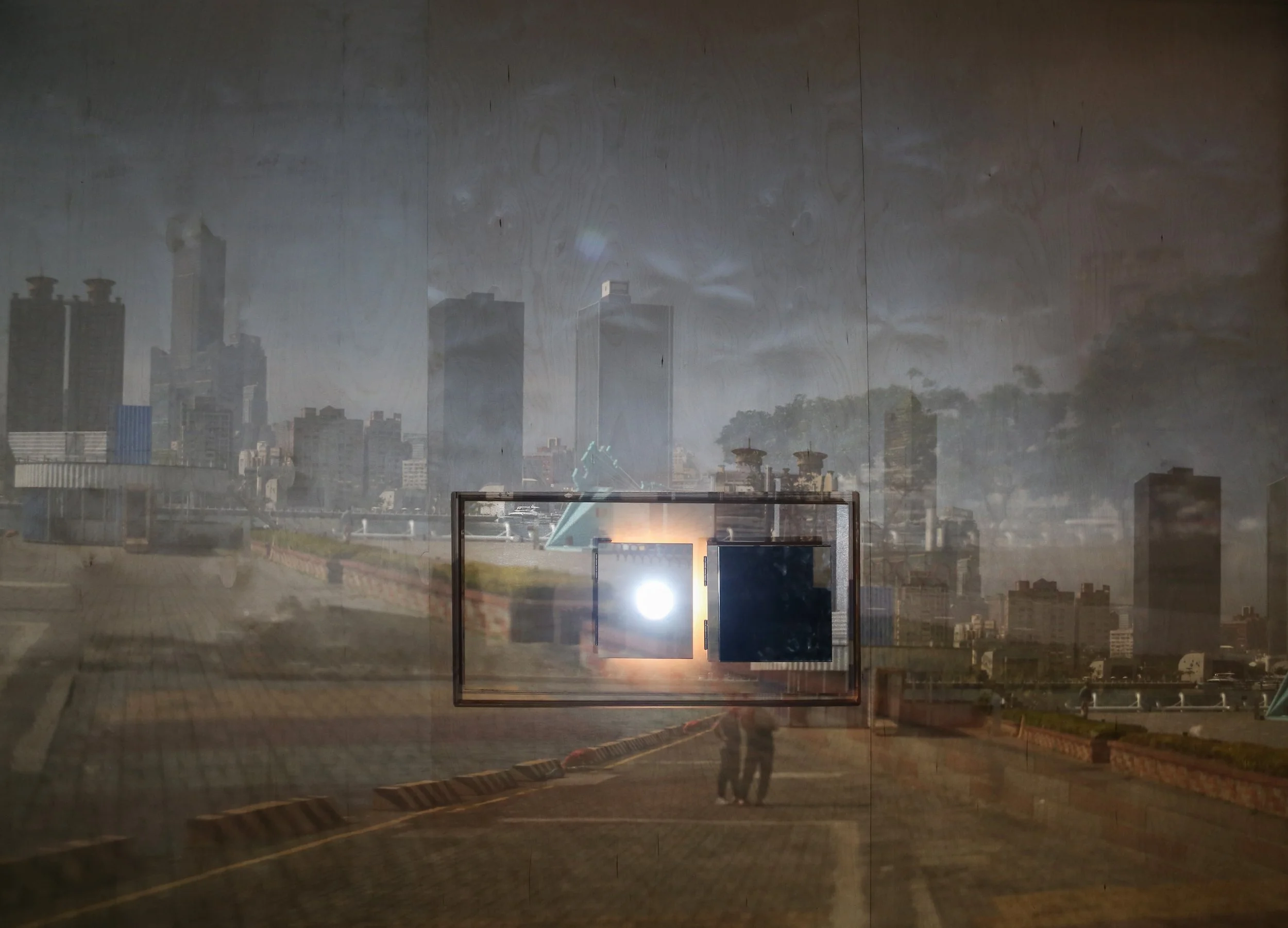 Image Description: The plywood wall with two projections of the Kaohsiung skyline overlapping. In the projections, two people are seen standing on the pier midway towards another container arts project. Many elements are projected twice. By Troels