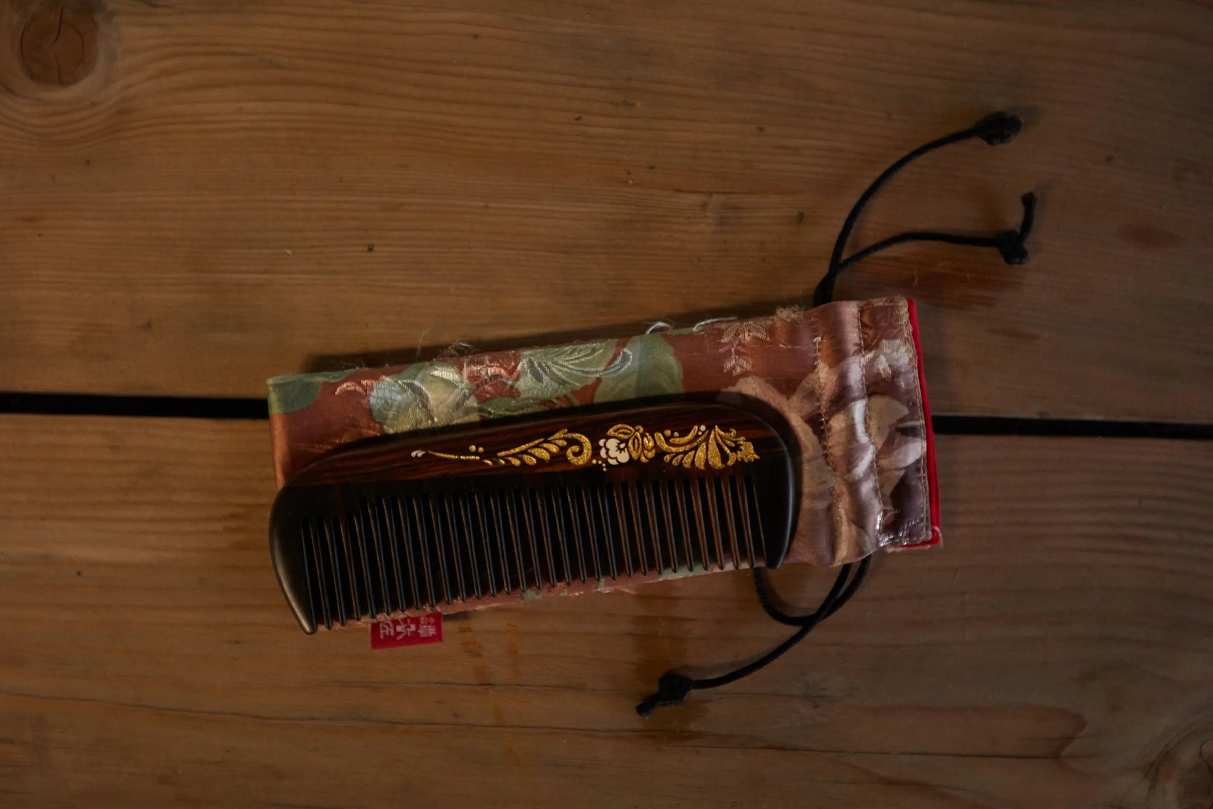 A close up of a dark tortoise comb with gold flower and pattern inscription laying on a dark pink fabric pouch with green and whitish flowers on it, on a wooden table.