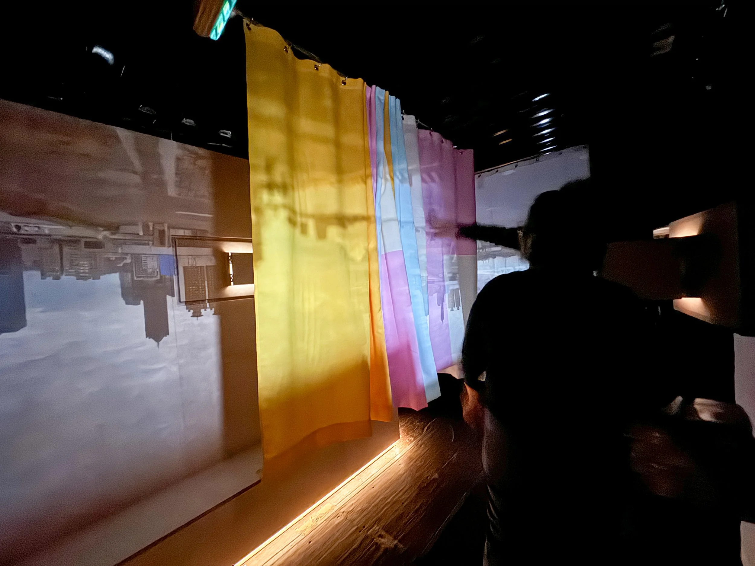 Image Description: Interior view of the Container Camera, where audiences are interacting with the work. Projections are seen on a large multi colored screen, a white screen, and a plywood wall. By Troels Steenholdt Heiredal