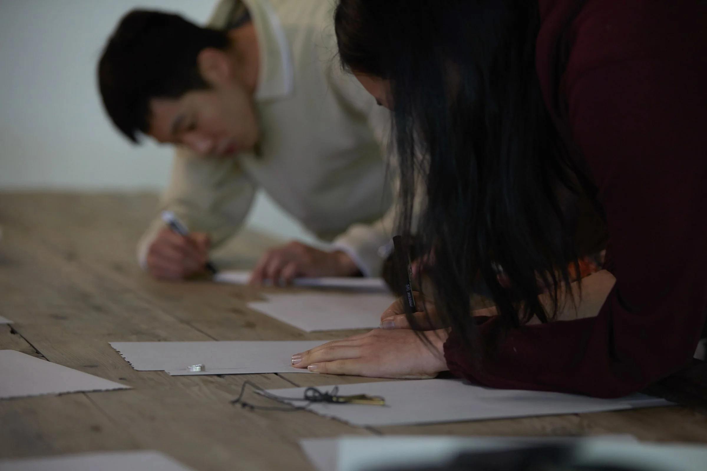 Close-up of a person in the foreground, mostly their long black hair visible, and hands on a large wooden table, writing one of several pieces of paper scattered with small objects on them. In the background, another person writing.