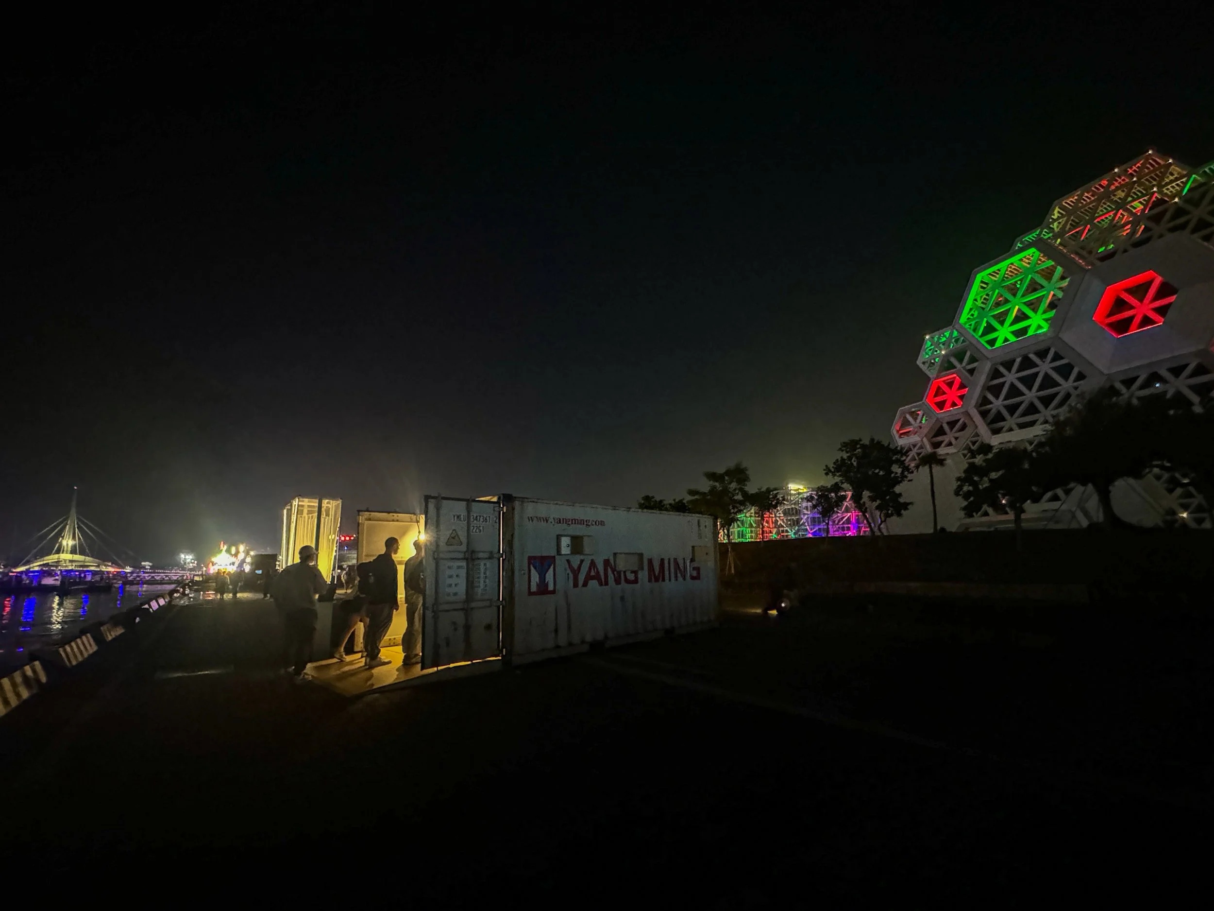 Exterior view of people waiting to get into the Container Camera, light comes from the opening, in the background are other container arts projects lit up, along with the bridge and the Pop Music Center.