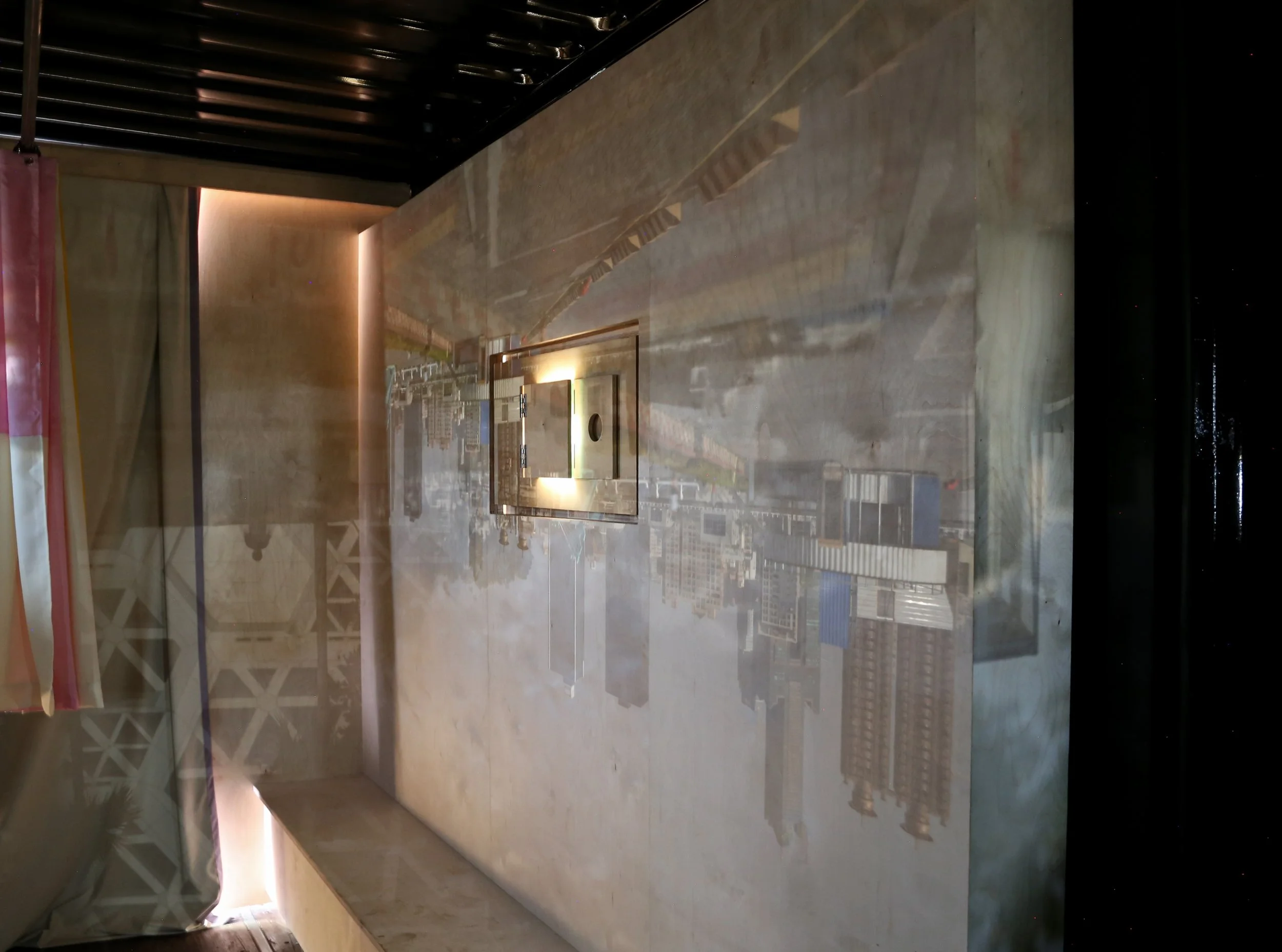 Image Description: Interior view of the Container Camera, showing the plywood wall and built-in bench. A lens box penetrates the wall, upon which two projections of the surroundings overlap.