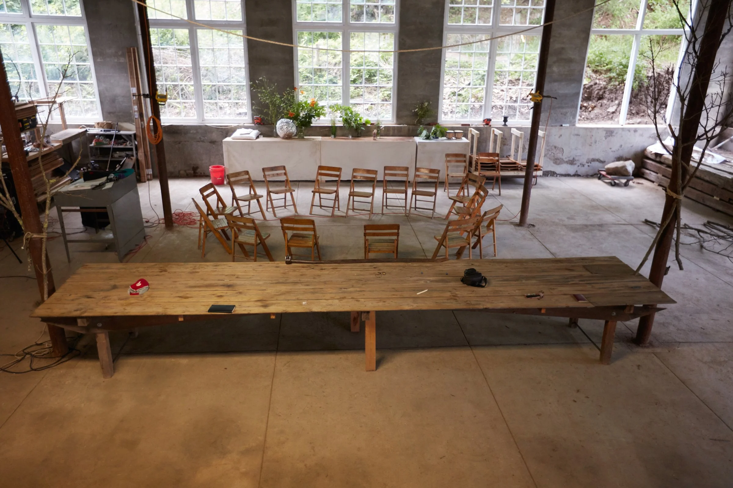 an historic industrial space, with large windows in the back, in the front a large table scattered with eight visible objects and behind it a circle of empty wooden chairs.