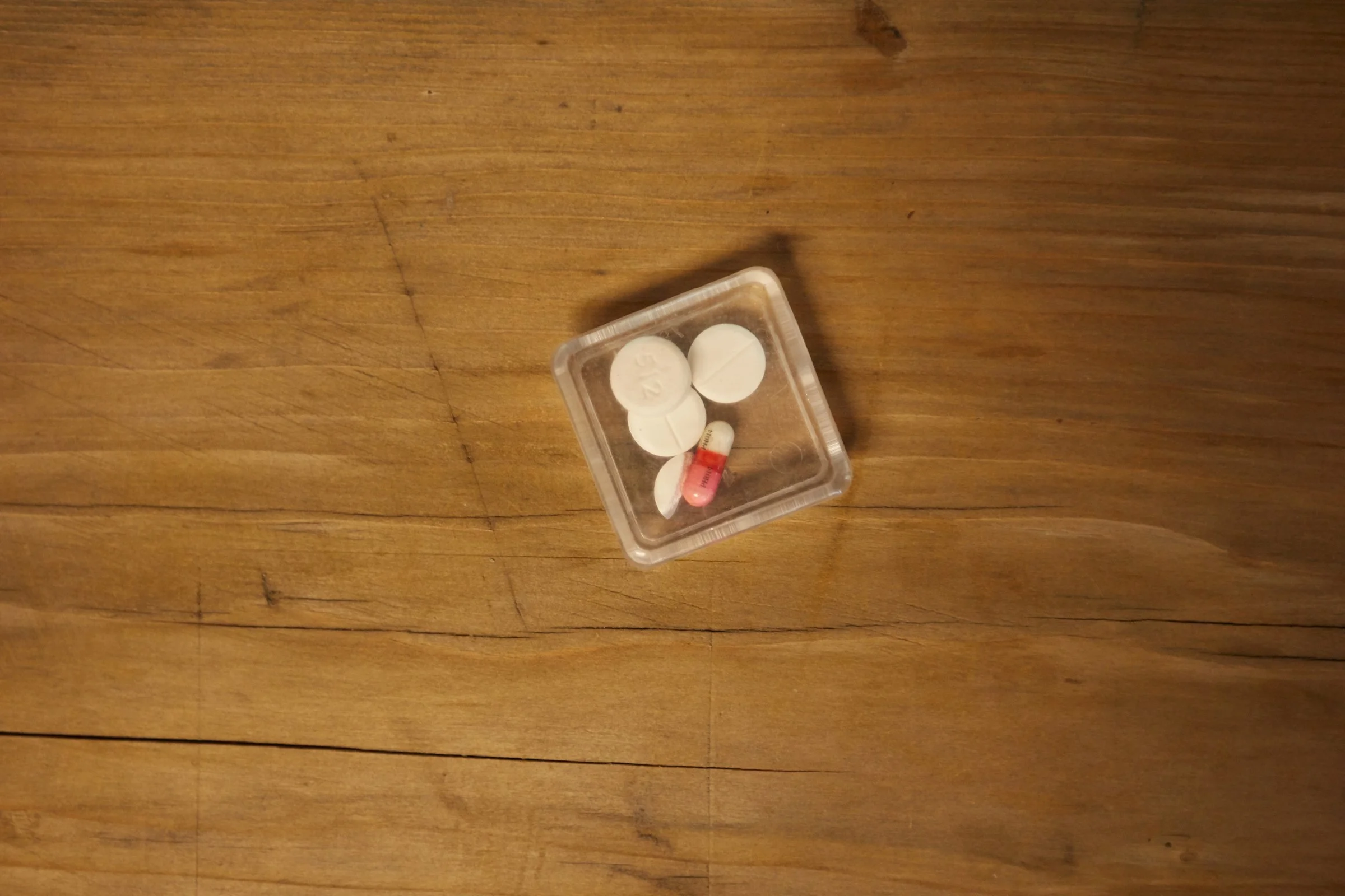 A close up of a small clear square plastic container with four and a half pills in it, one white and pink, the rest all white.