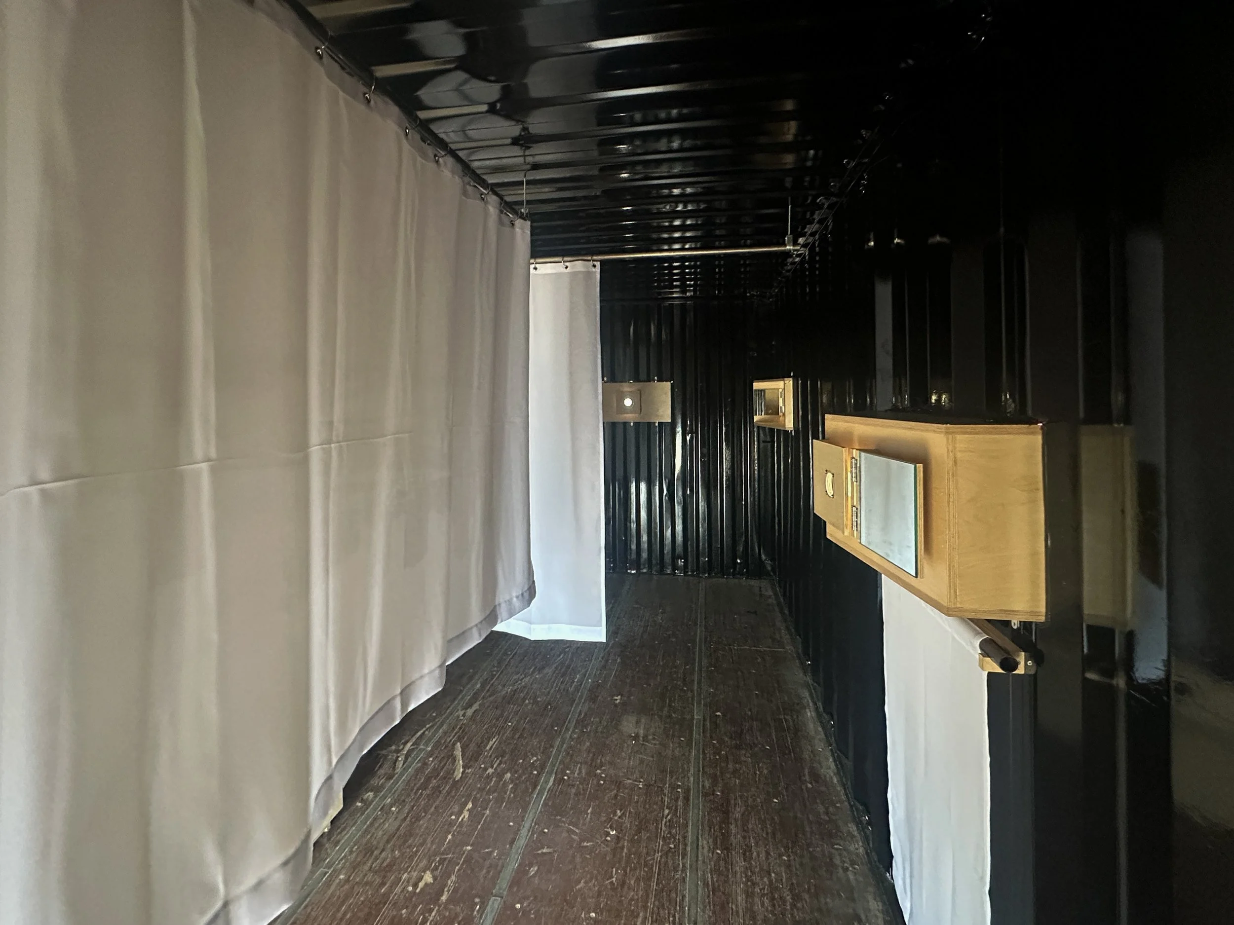 Image Description: Interior view of the Container Camera, showing the plywood lens boxes through the container wall and the screens hanging in the center of the space. By Autistic architect and artist, Troels Steenholdt Heiredal