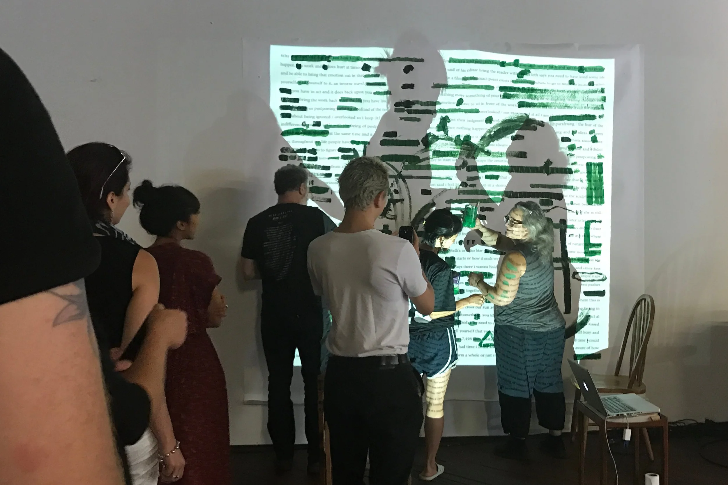 A line of people waiting to get up to a poem projected onto a large sheet of paper on the wall, where they will paint over words with green paint, a person is painting over words on the arm of another person.