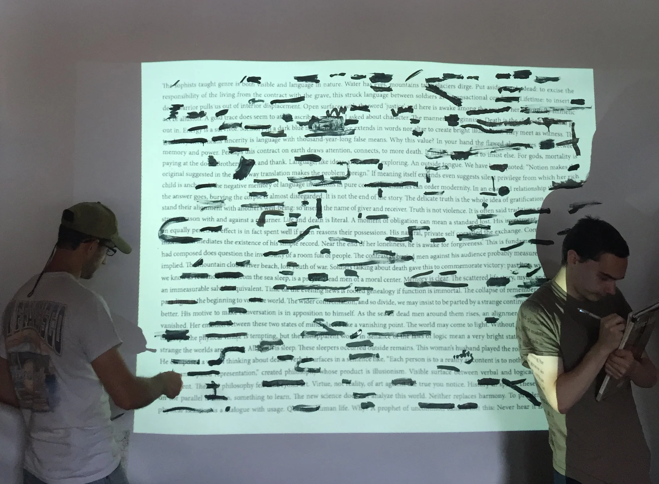 Photo of a poem projected onto a large sheet of paper. Many words are painted over with green paint. On either side stands a person, the left is painting on the poem, the right is writing in their notebook.