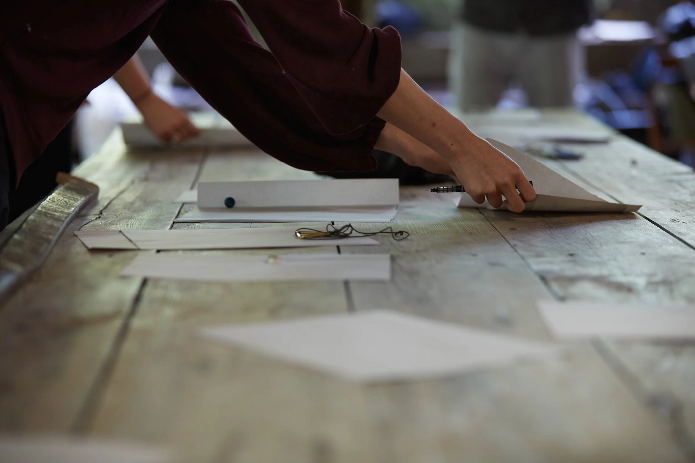 Photo of a large wooden table with parts scattered over it. Tow hands are in the process of lifting a piece of paper off the table while also holding a pen.