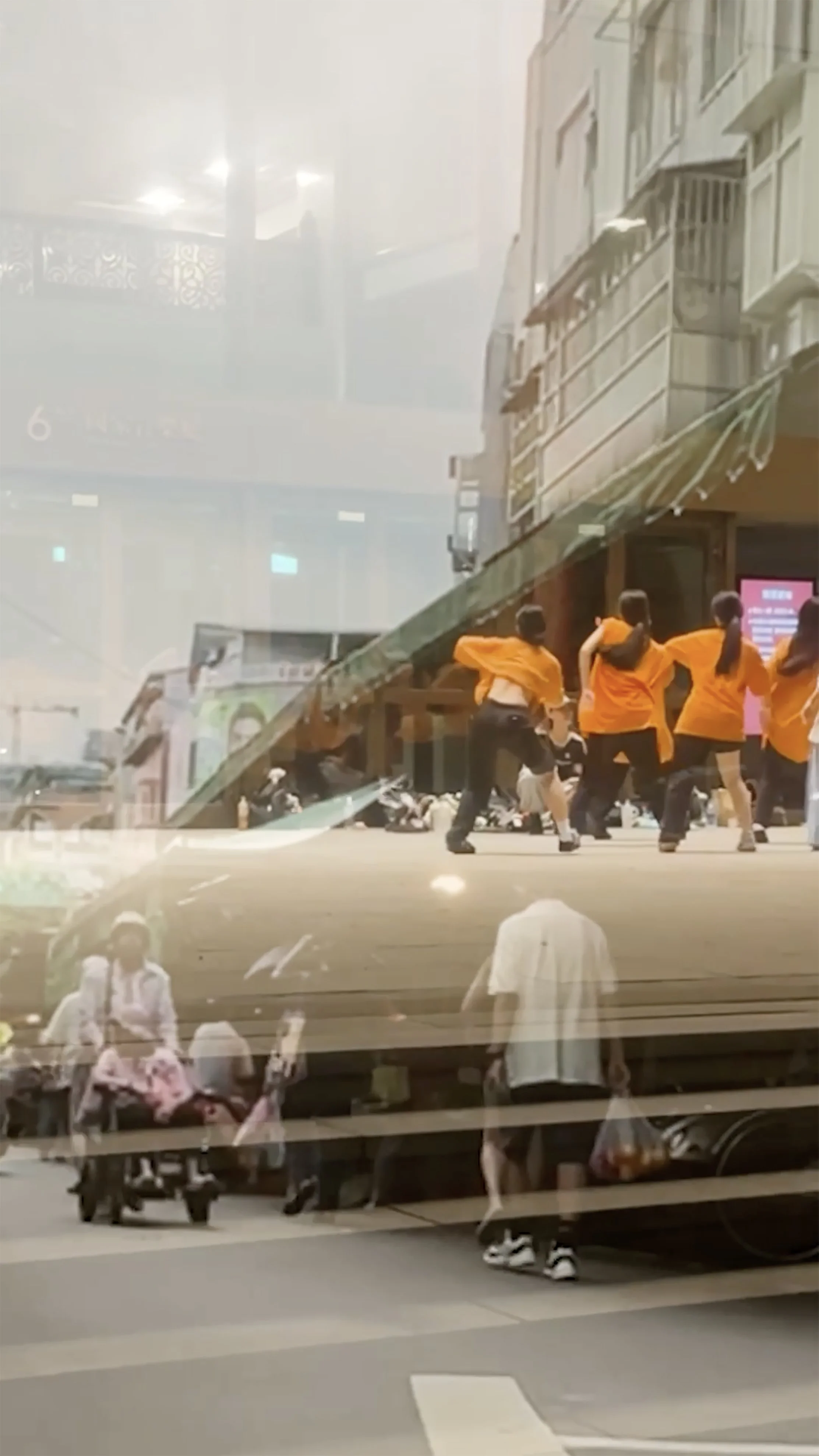 Screenshot from the film. Two scenes are merging: one showing people shopping at an outdoor wet market, the other of young people in black pants and oragne baggy t-shirts dancing in front of a glazed storefront, using it as a mirror