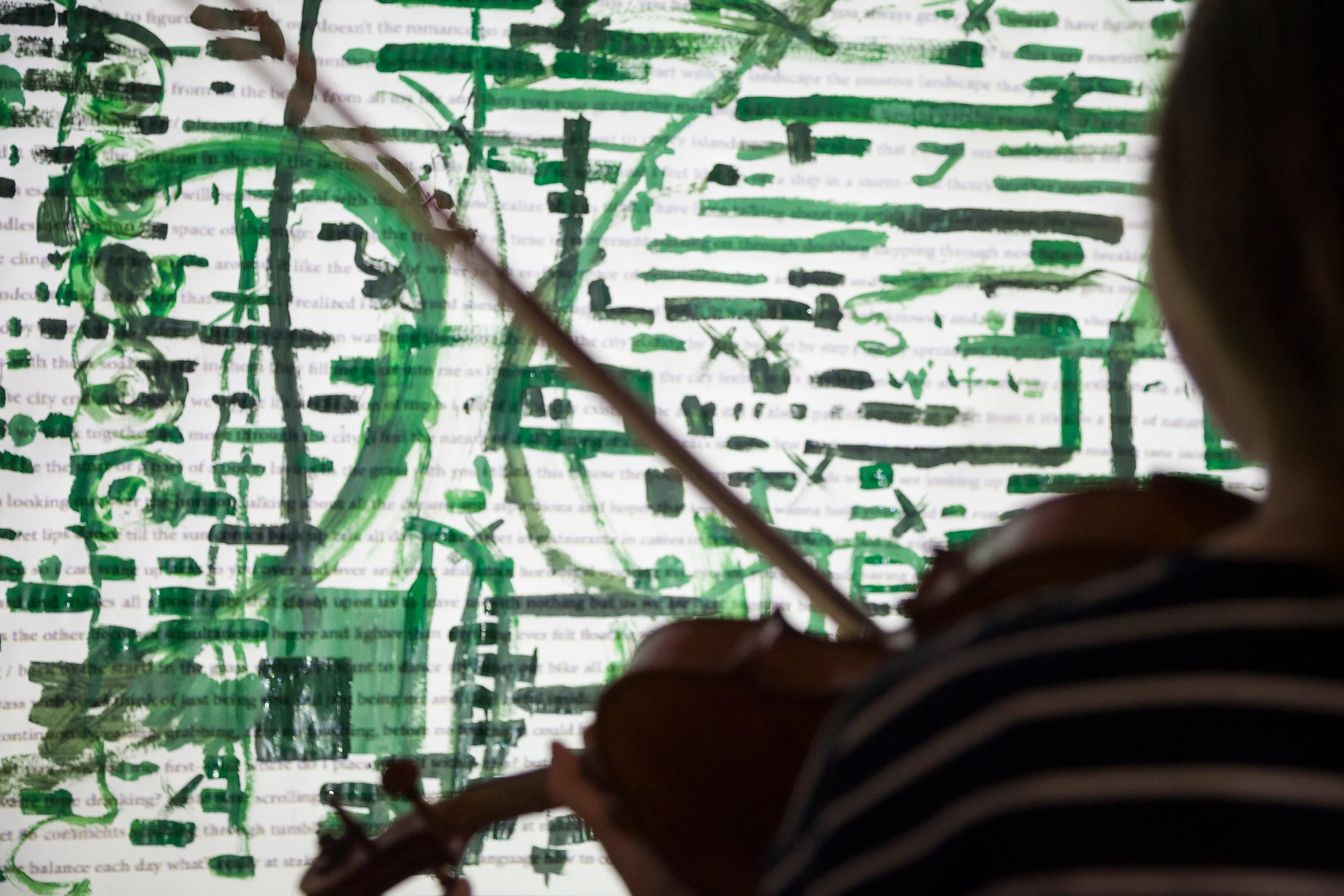 Looking over the shoulder of a violinist at a wall where a texts if projects and covered in green painting marks.