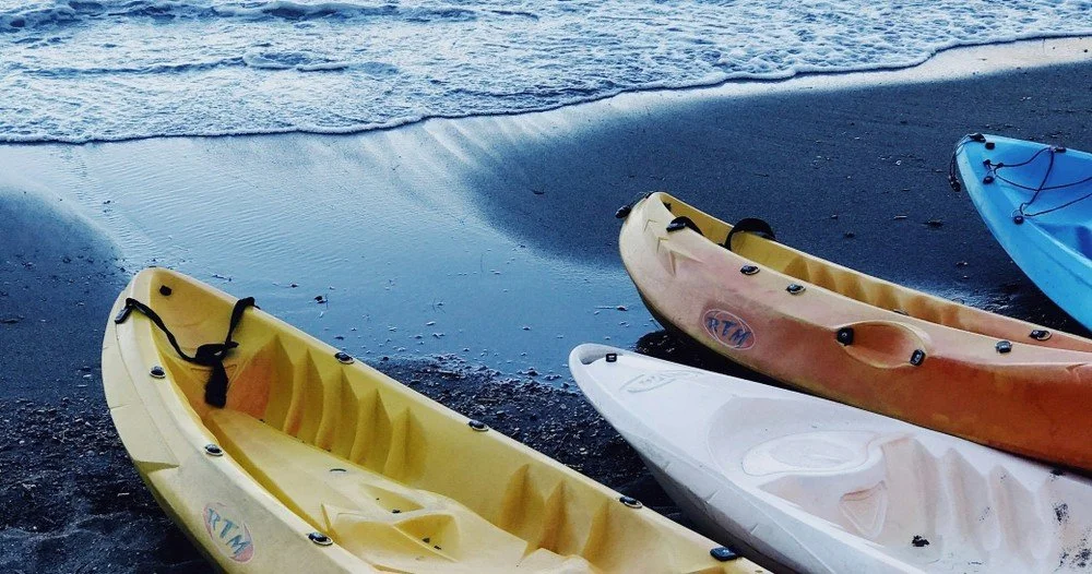 Two people kayaking on open water, with one person wearing a black cap and the other with hair tied up, both sitting in an orange kayak.