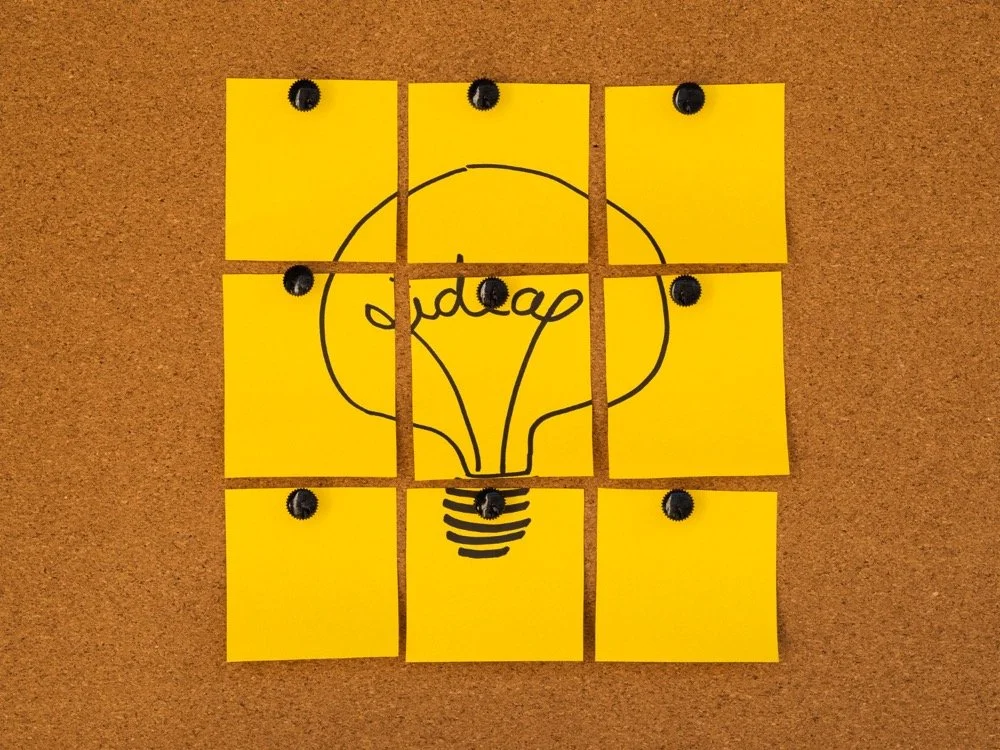 A corkboard with nine yellow squares attached with screws, arranged in a 3x3 grid. The middle square features a black outline of a light bulb with the word 'idea' written inside it. The screws are in the top left and right corners of the top row, the middle left and right of the middle row, and the bottom left and right corners of the bottom row.