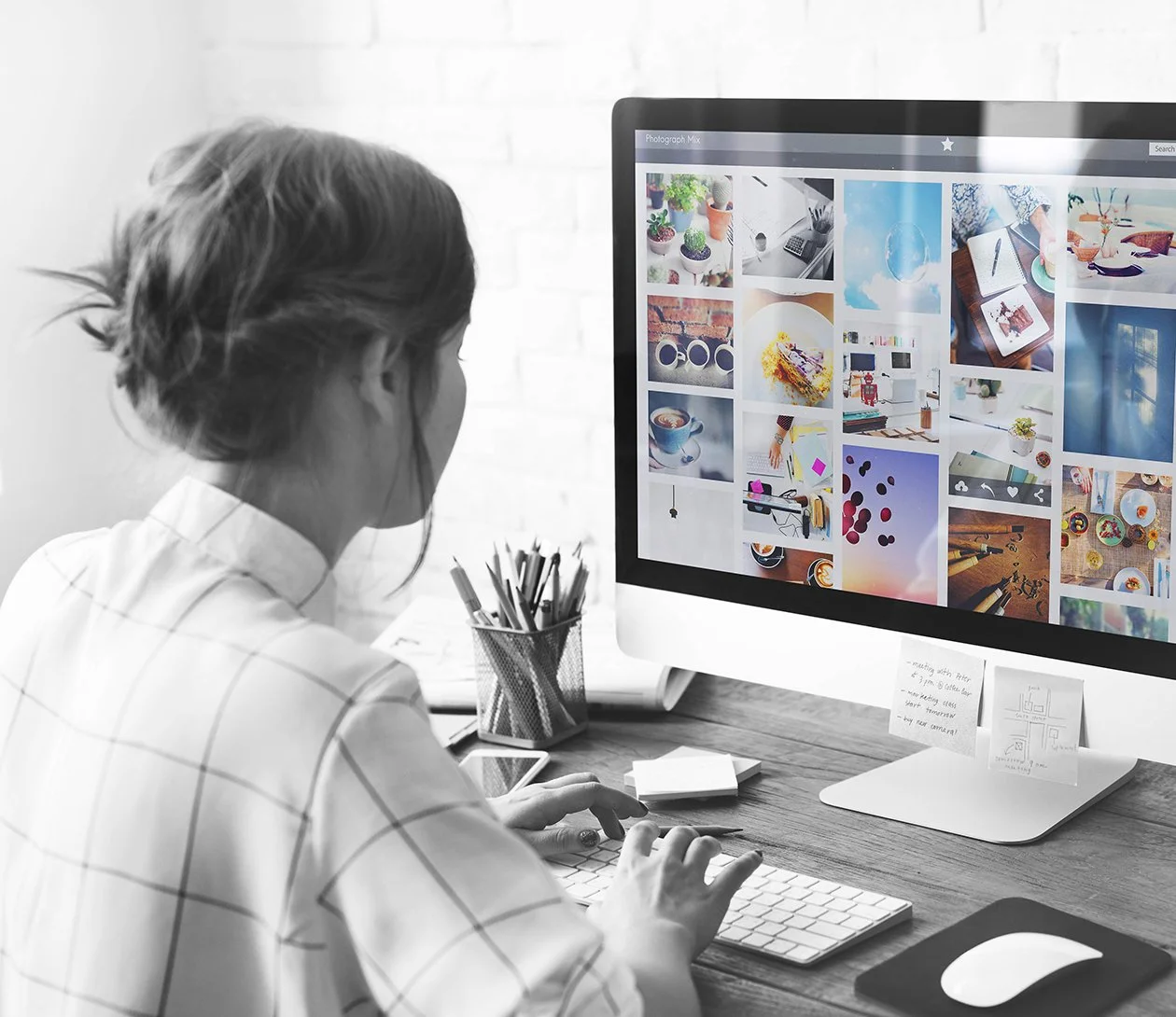A woman working at a desk with a large desktop computer displaying a collage of colorful images, surrounded by stationery and a smartphone.