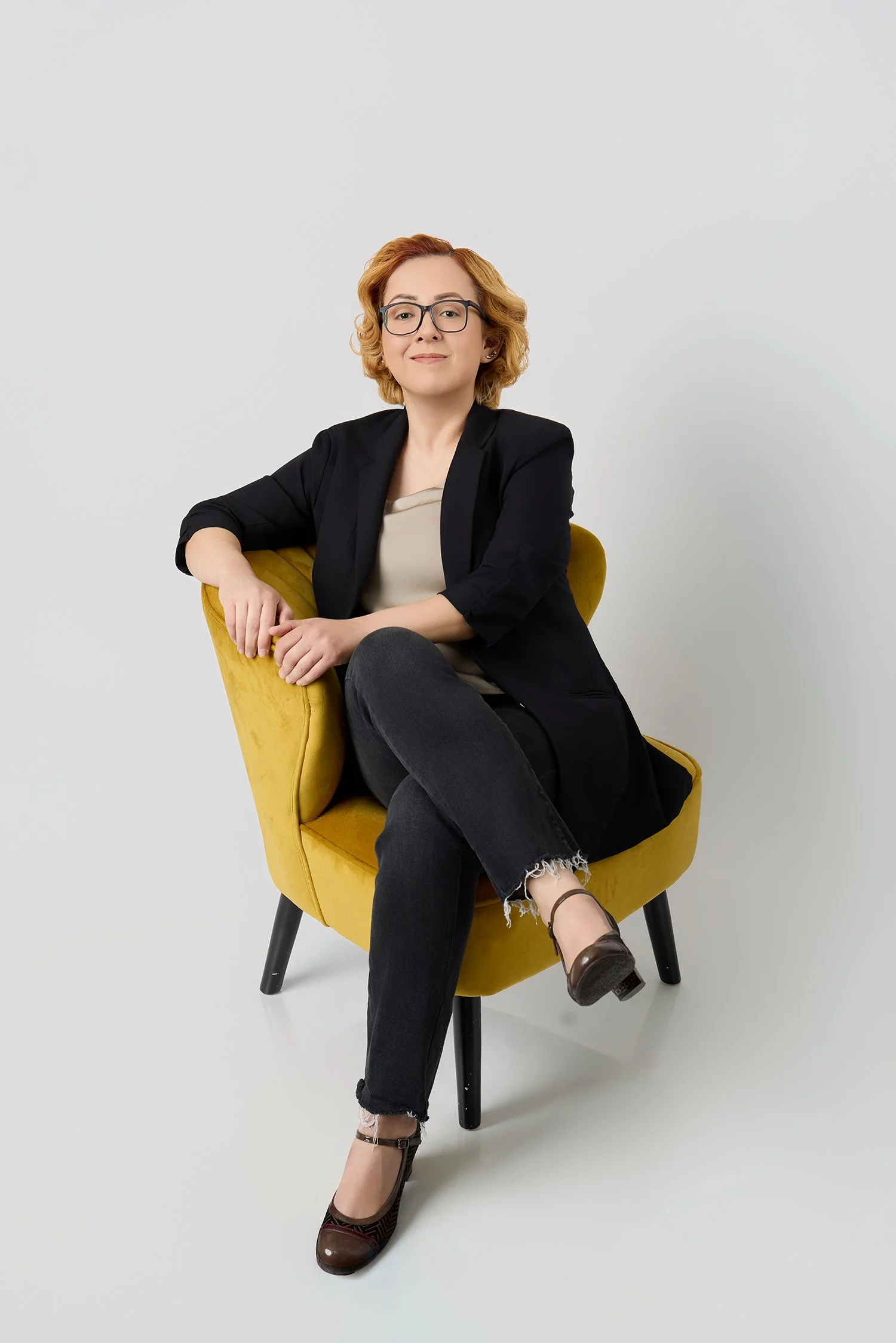 A woman with short red hair, glasses, and a black blazer sitting on a yellow chair against a plain white background.