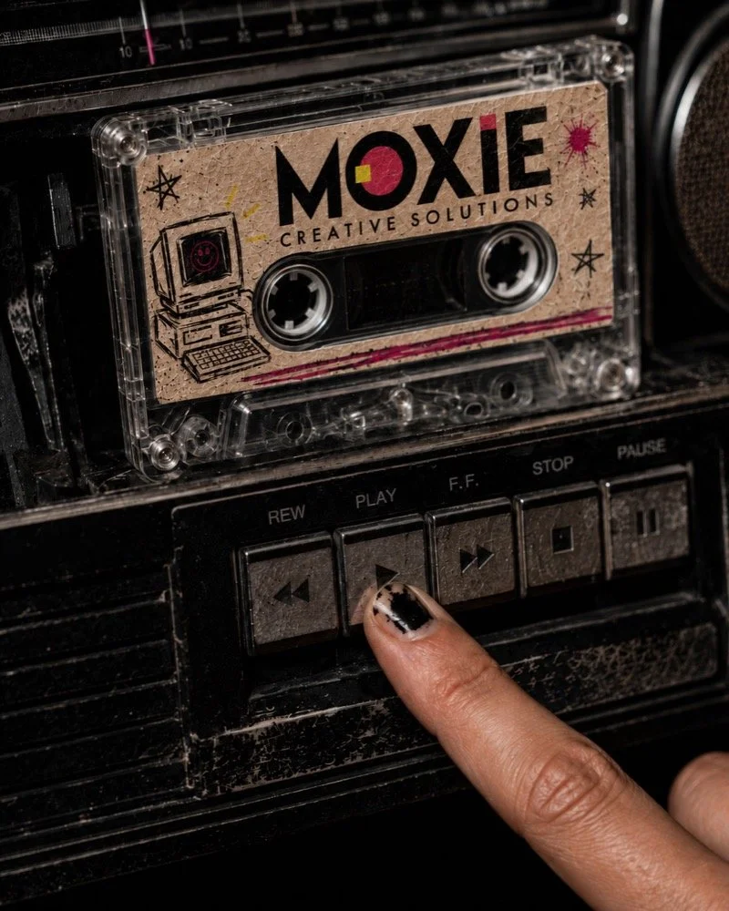 A person's finger with chipped black and white nail polish pressing a button on a vintage cassette tape player, with a cassette tape labeled 'Moxie Creative Solutions' inserted in the player.