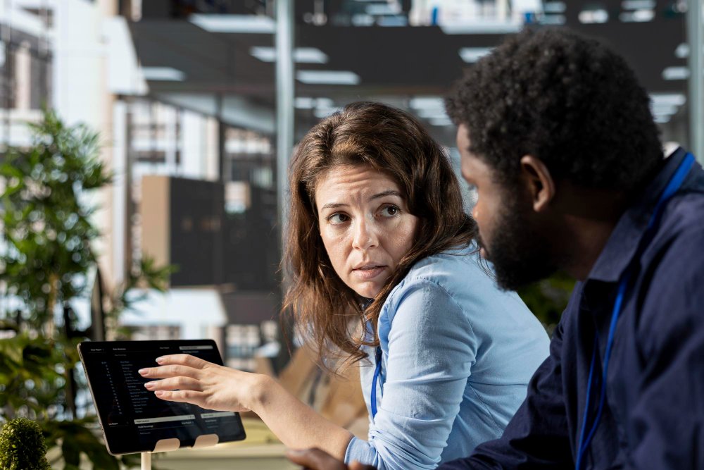 A woman pointing at a digital tablet while discussing business data and messaging constraints with a colleague.