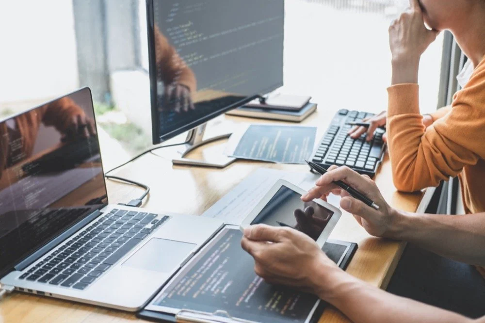 Two people working at a desk with multiple screens, laptops, a tablet, and notes, focusing on coding or programming.