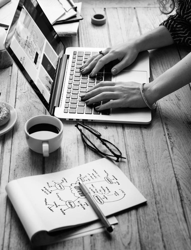 Person typing on a laptop at a wooden desk with notebooks, glasses, a cup of coffee, a plate with a snack, and stationery.
