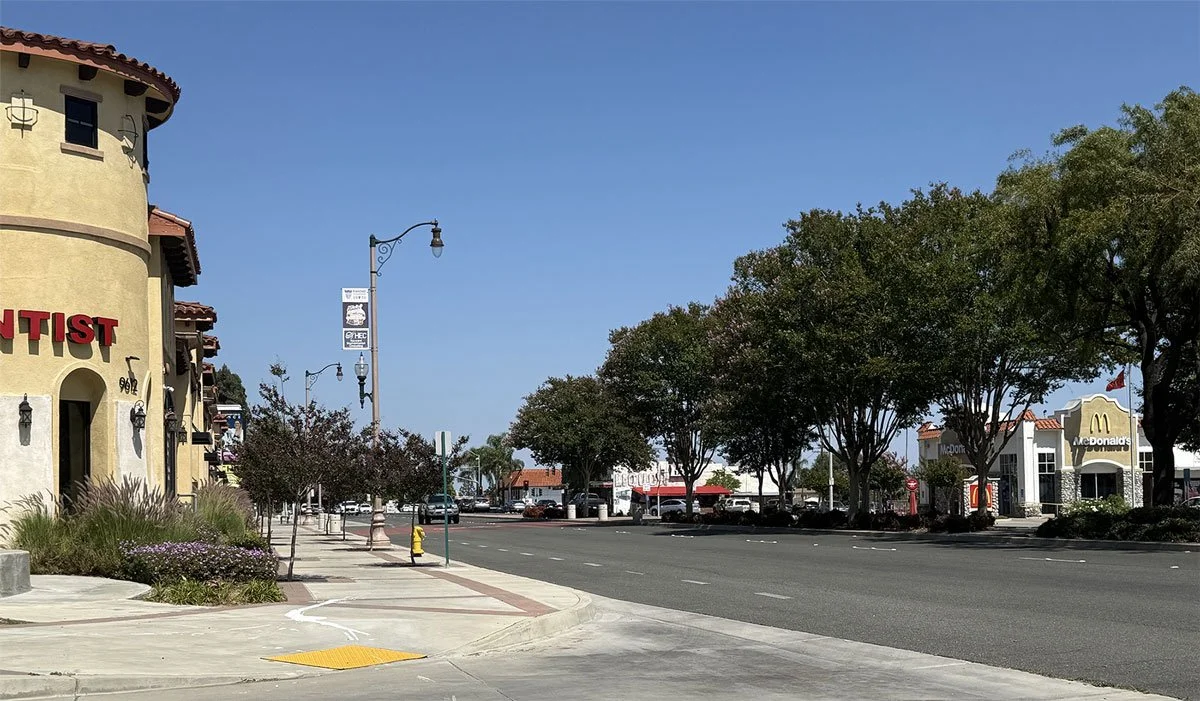 A quiet street scene with McDonald's restaurant on the right, an apartment building with a painted sign on the left, trees lining the sidewalk, parked cars on the street, and clear blue sky overhead.