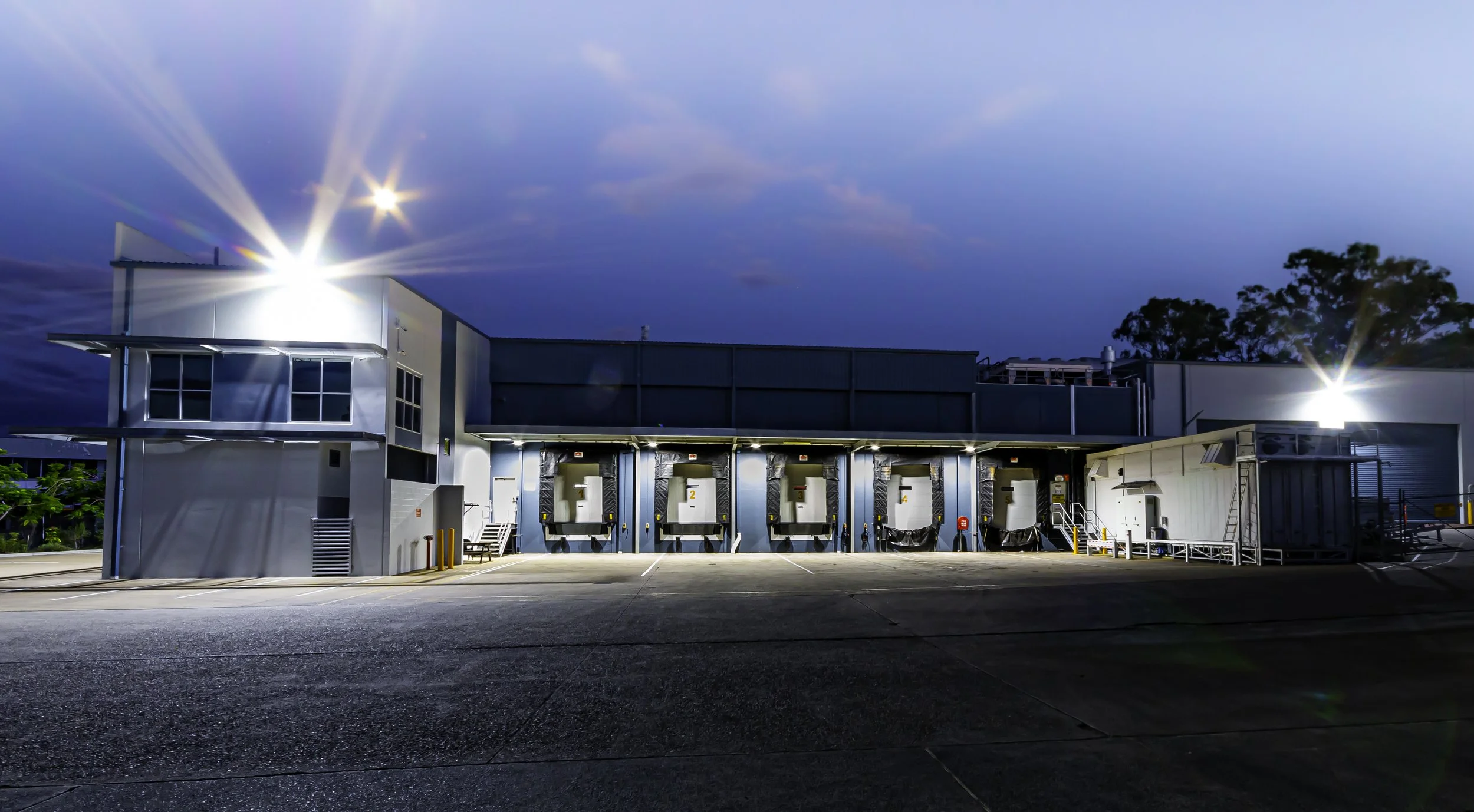 Late evening outside view of a modern industrial building with docking bays, bright exterior lights, and an empty paved area in front. The sky is dark blue with clouds, and there are trees in the background.