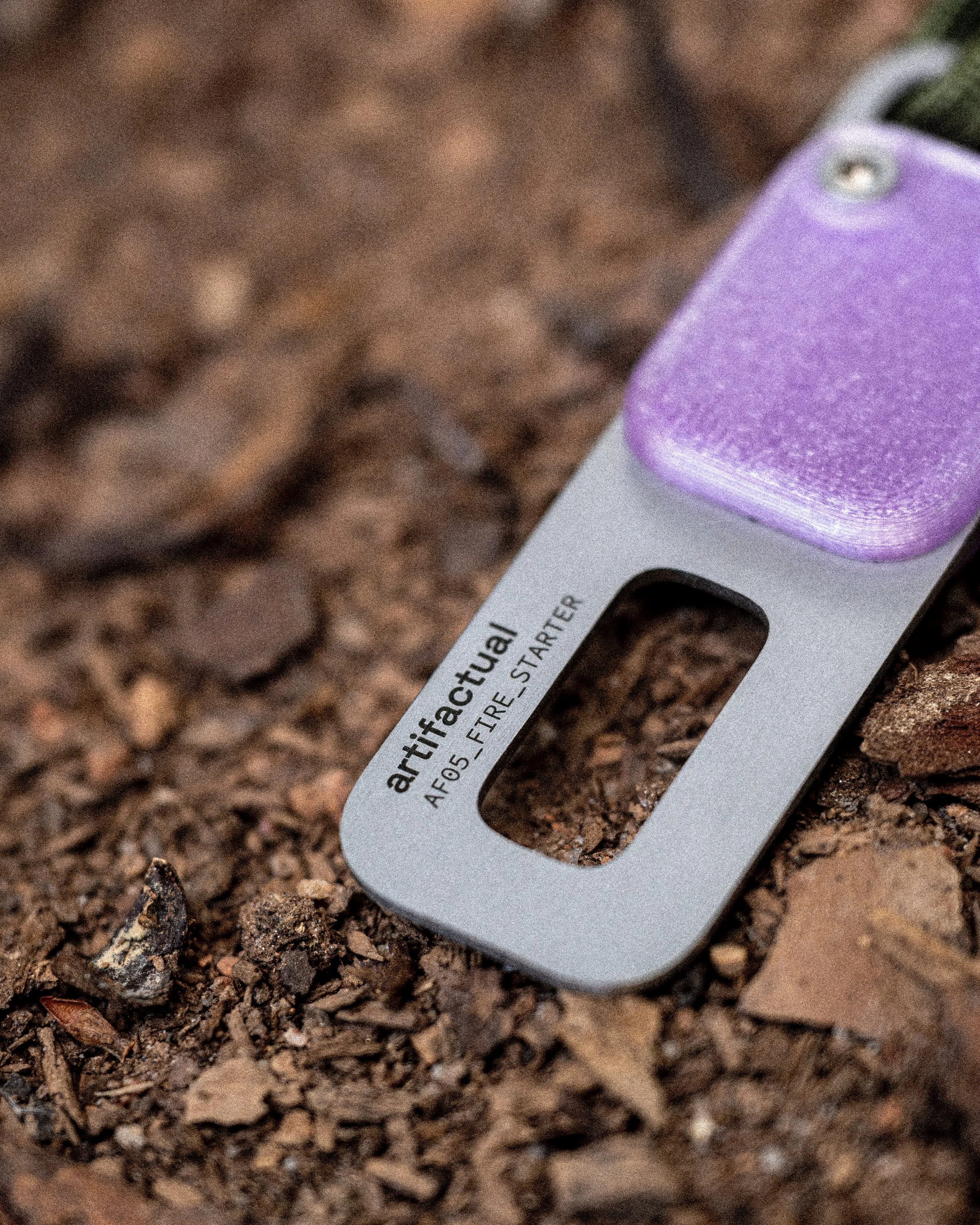 Close-up of a fire starter and ferro-rod kit lying on the ground covered with dirt and small rocks, with a purple fire striker tool visible.