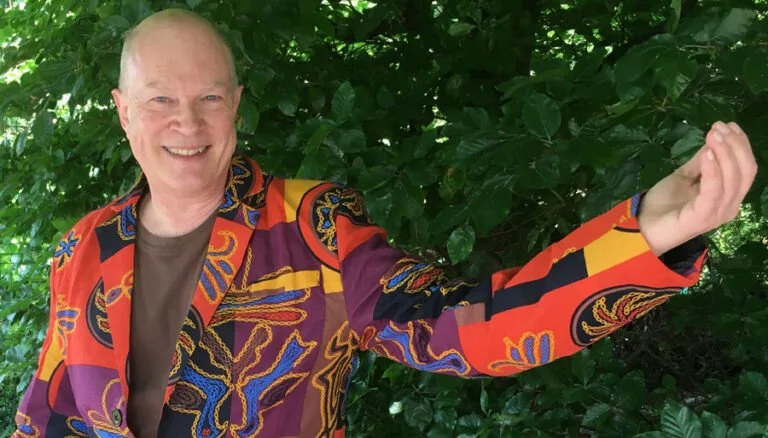 A smiling man wearing a colorful patterned blazer extending his arm in front of green foliage.