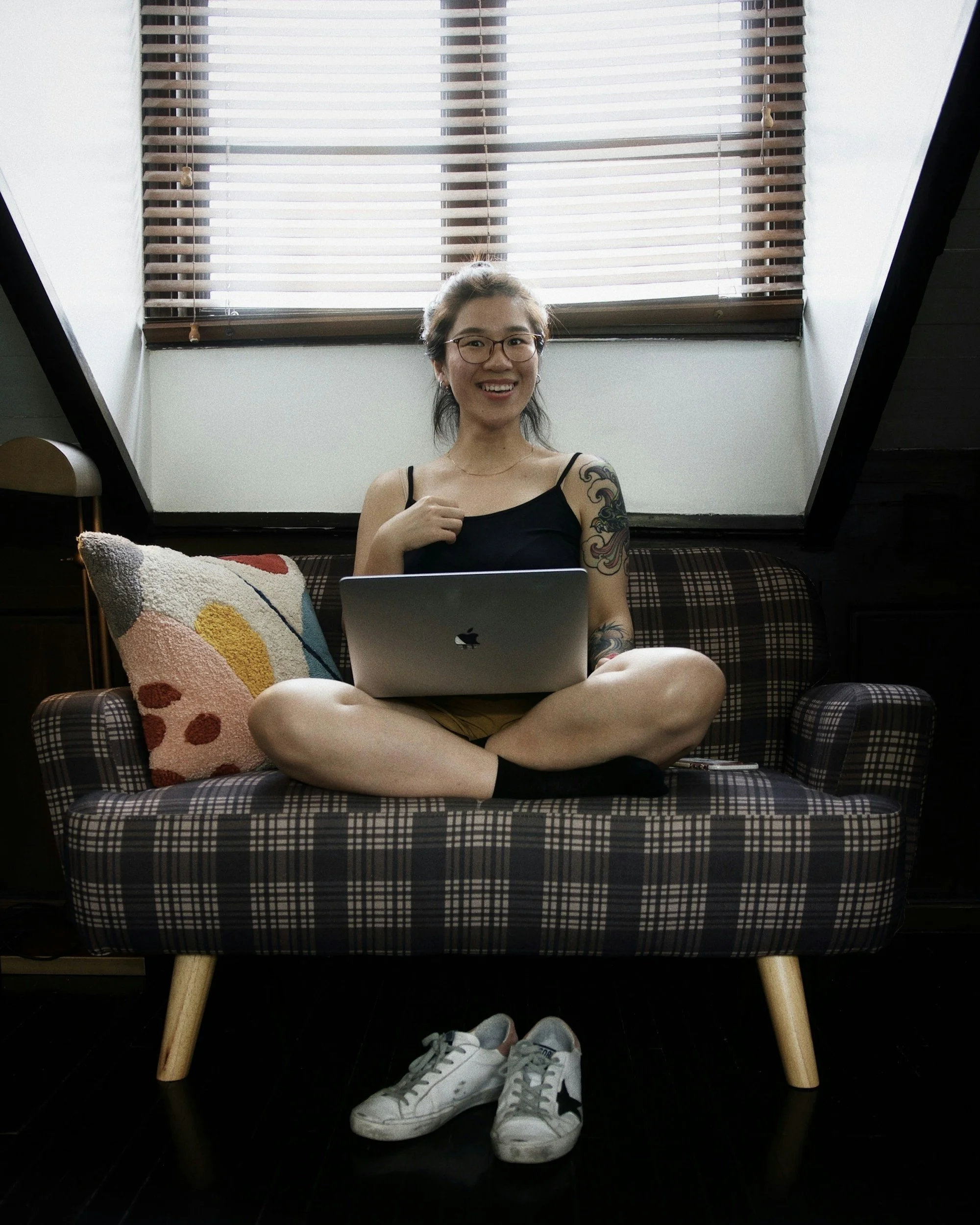 A young woman with glasses and tattoos sitting cross-legged on a plaid sofa in front of a large window with blinds, holding a MacBook, smiling at the camera, with a colorful pillow and a pair of white sneakers on the floor in front.