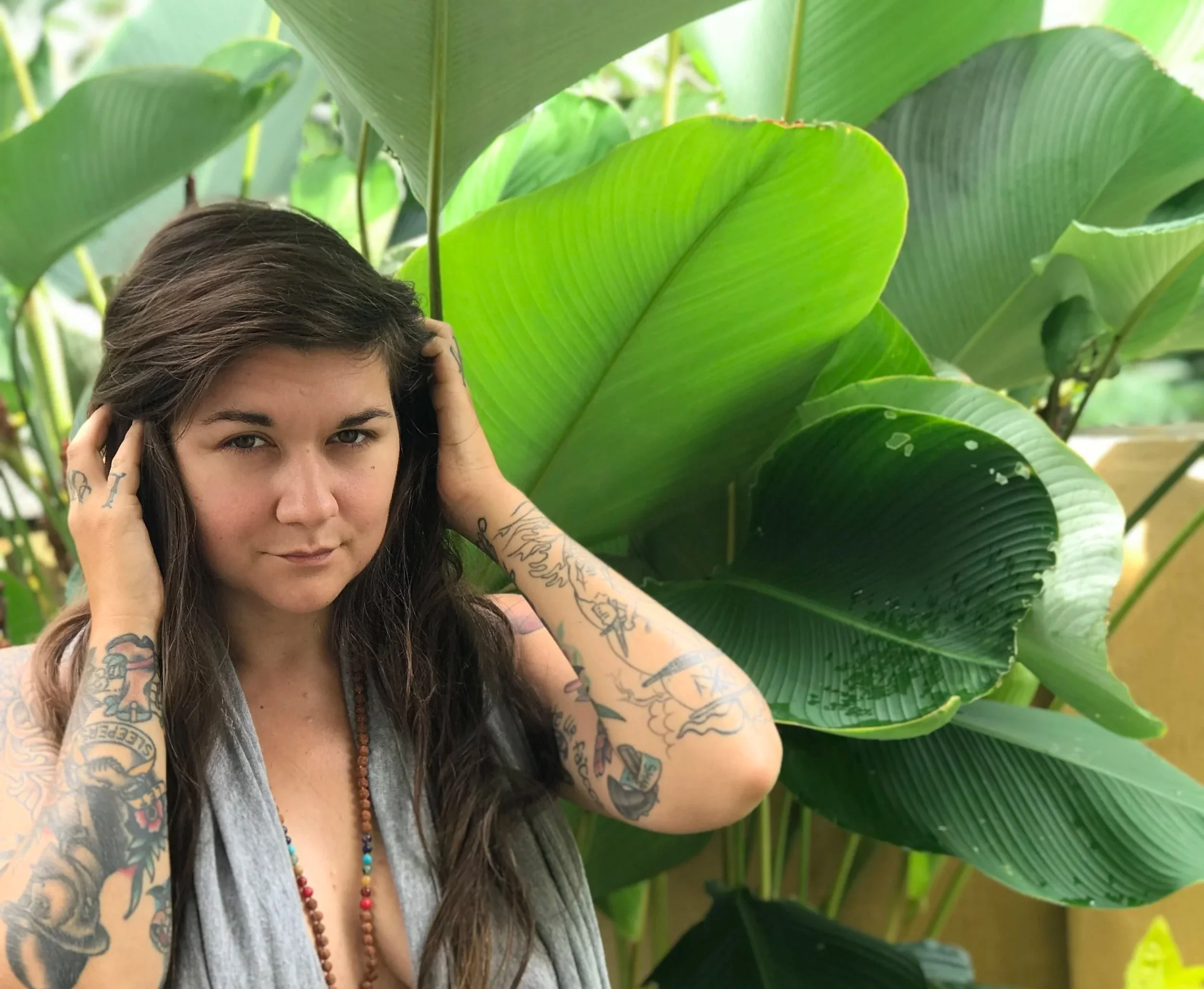 Young woman with long brown hair and tattoos on arms, holding her head with both hands, standing among large green tropical leaves.