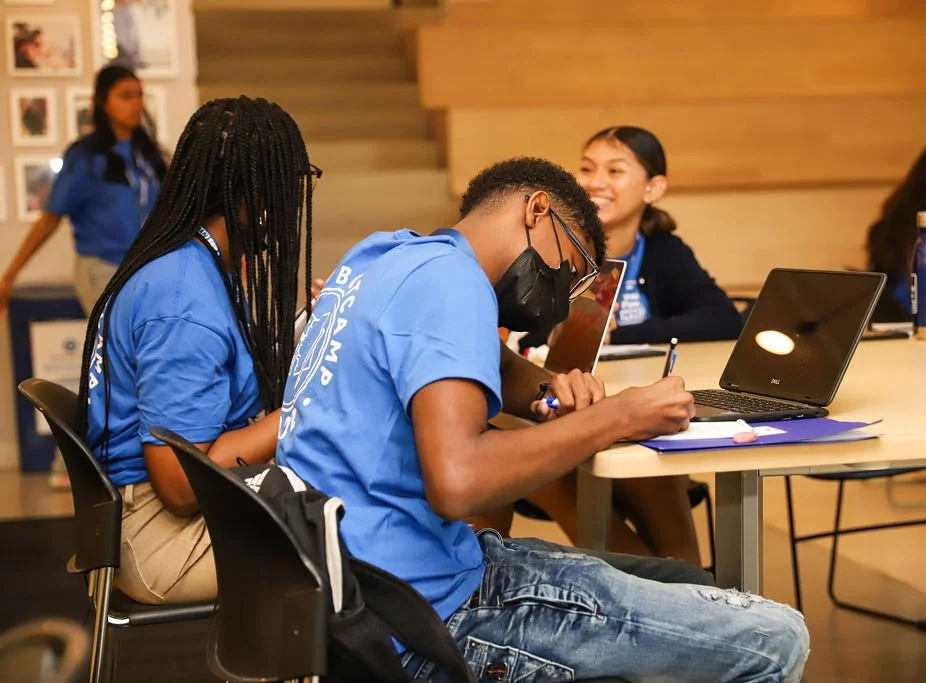 Group of young students in blue shirts sitting at tables with laptops and tablets, engaged in a classroom activity.