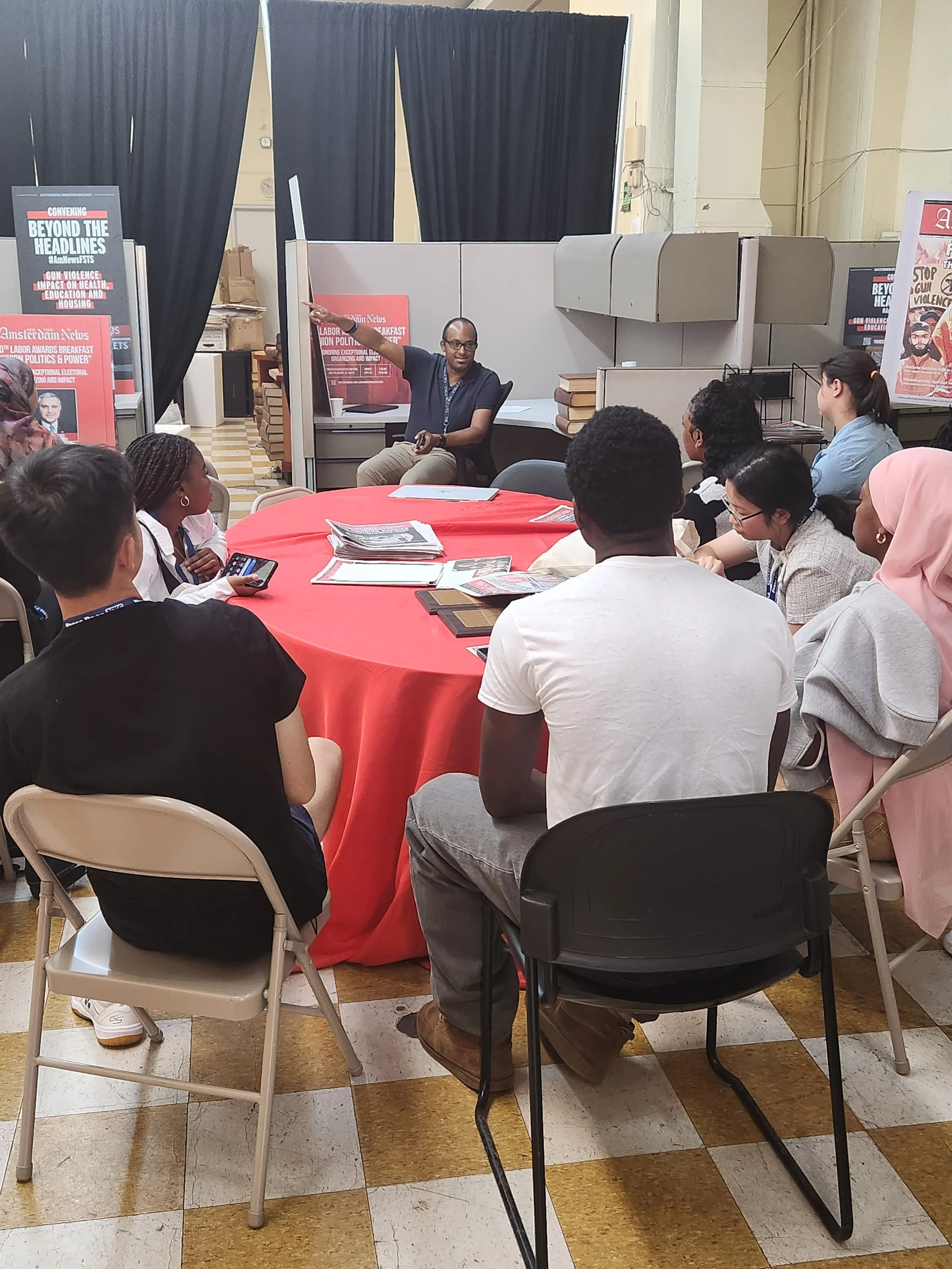 A man is speaking to a group of people seated around a red-covered round table, with posters in the background indicating a discussion or seminar on labor politics.