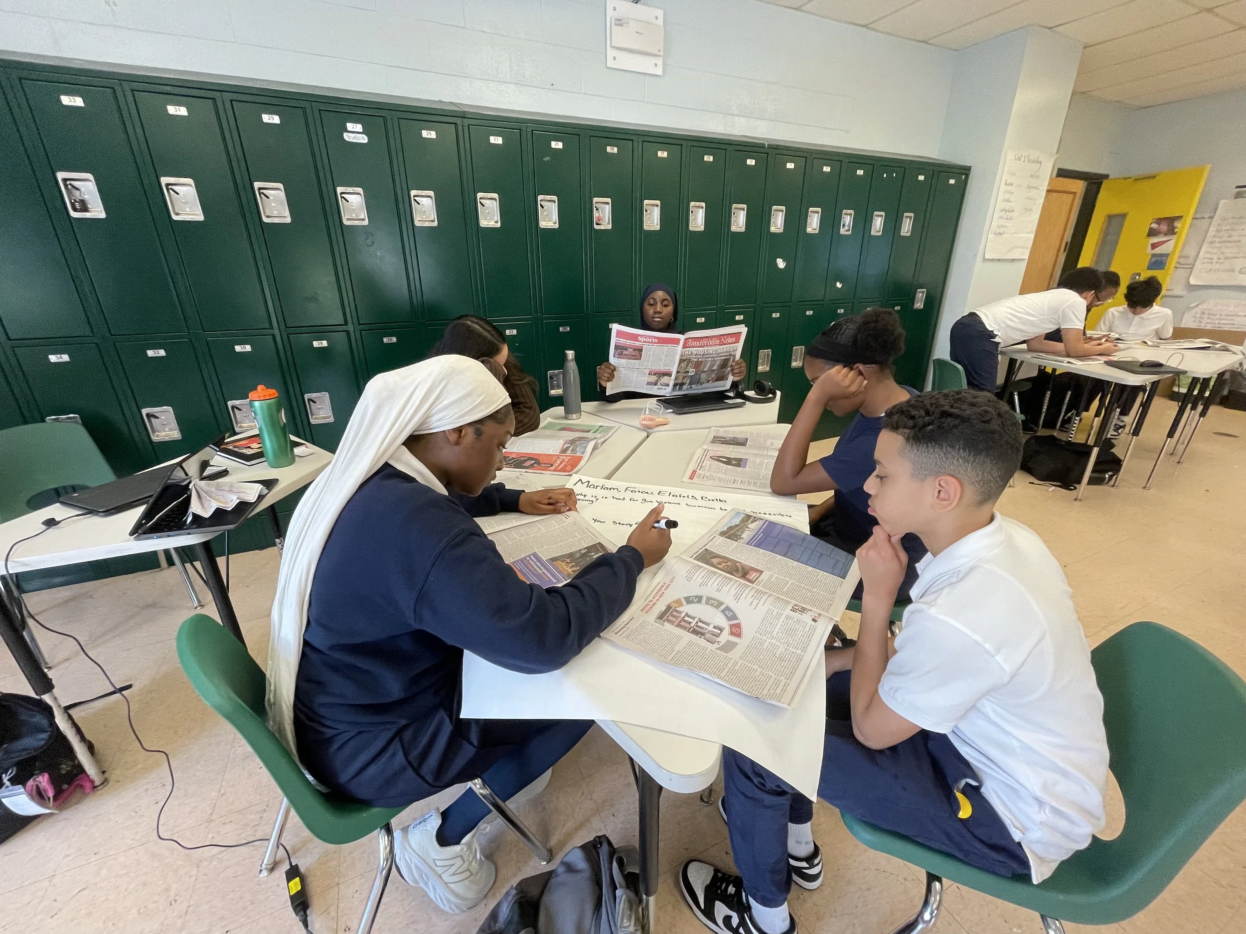 Students sitting at a table reading newspapers in a classroom with lockers in the background.
