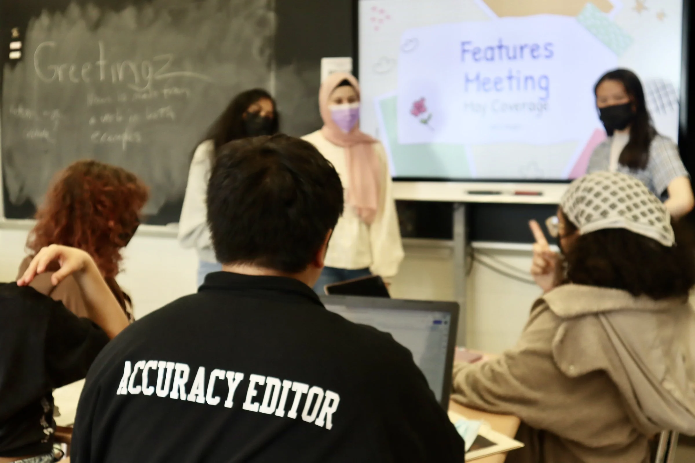 Classroom scene with five students, some wearing masks, listening to a presentation on a large screen that reads 'Features Meeting' and 'Hey Coverage.' A chalkboard in the background has writing that says 'Greeting.'