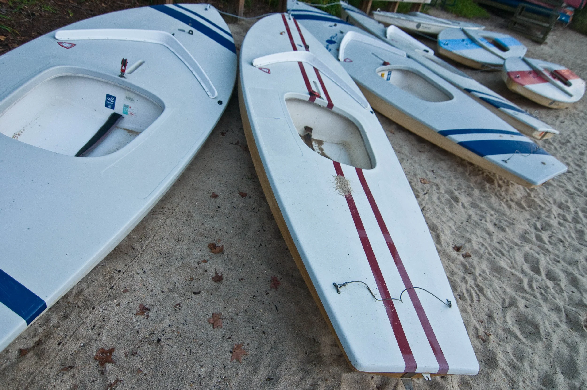 Several small sailboats with white hulls and colored stripes are lying on sandy ground outdoors. Some boats have red or blue stripes, and they are equipped with small anchors and rigging.